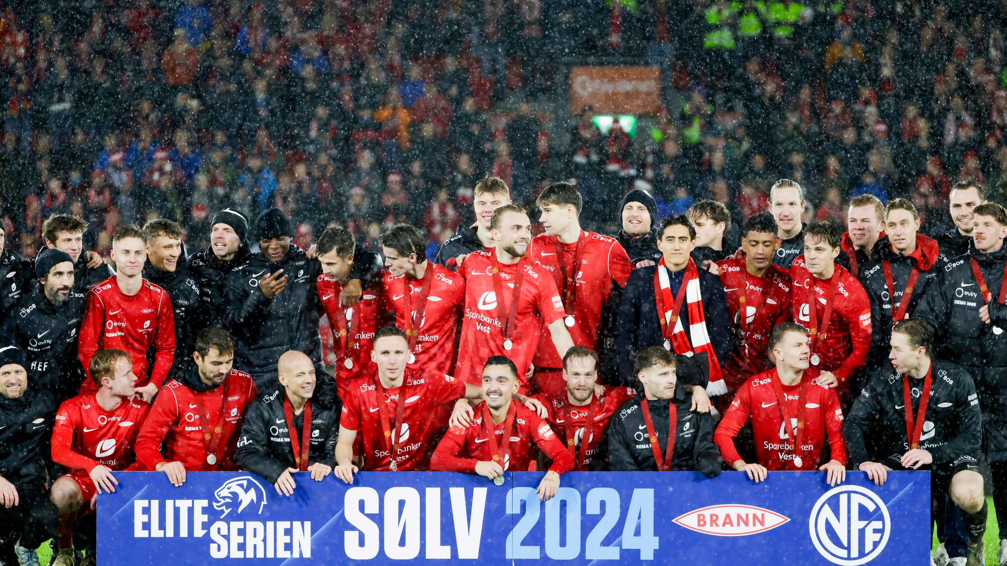 Spillere fra Brann feirer sølv i Eliteserien 2024 med medaljer rundt halsen på banen. (Bildebeskrivelsen er laget av en KI-tjeneste)