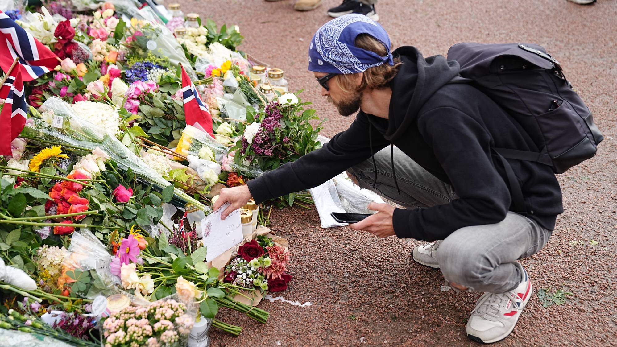 Person legger ned blomster og et kort ved et minnested med mange blomsterbuketter og norske flagg. (Bildebeskrivelsen er laget av en KI-tjeneste)