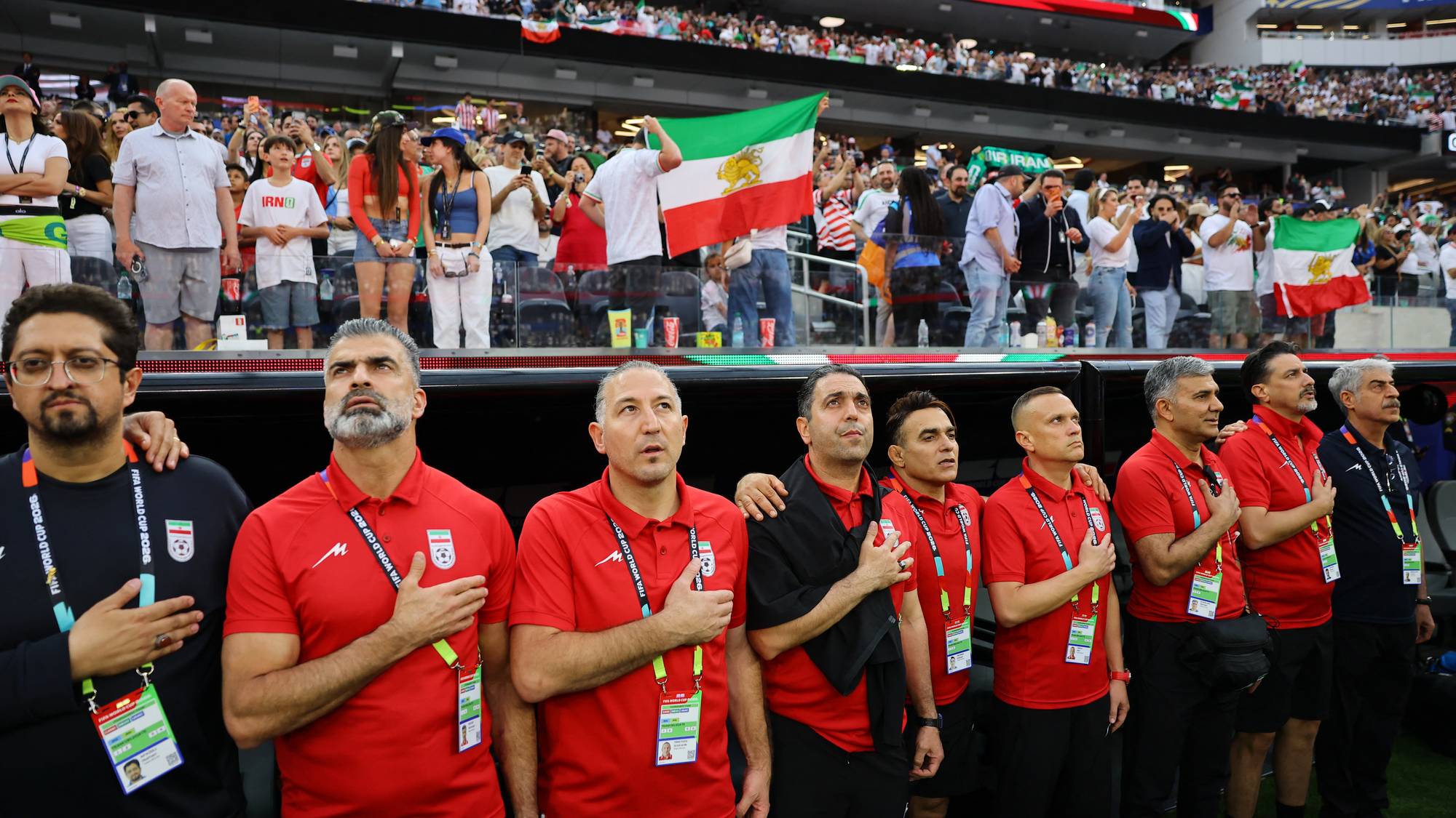 En gruppe menn i røde overtrekksgensere står med hånden på brystet under en fotballkamp. Bak dem står tilskuere på en tribune med iranske flagg. Mannen til venstre bærer briller og har på seg en svart genser. (Bildebeskrivelsen er laget av en KI-tjeneste)