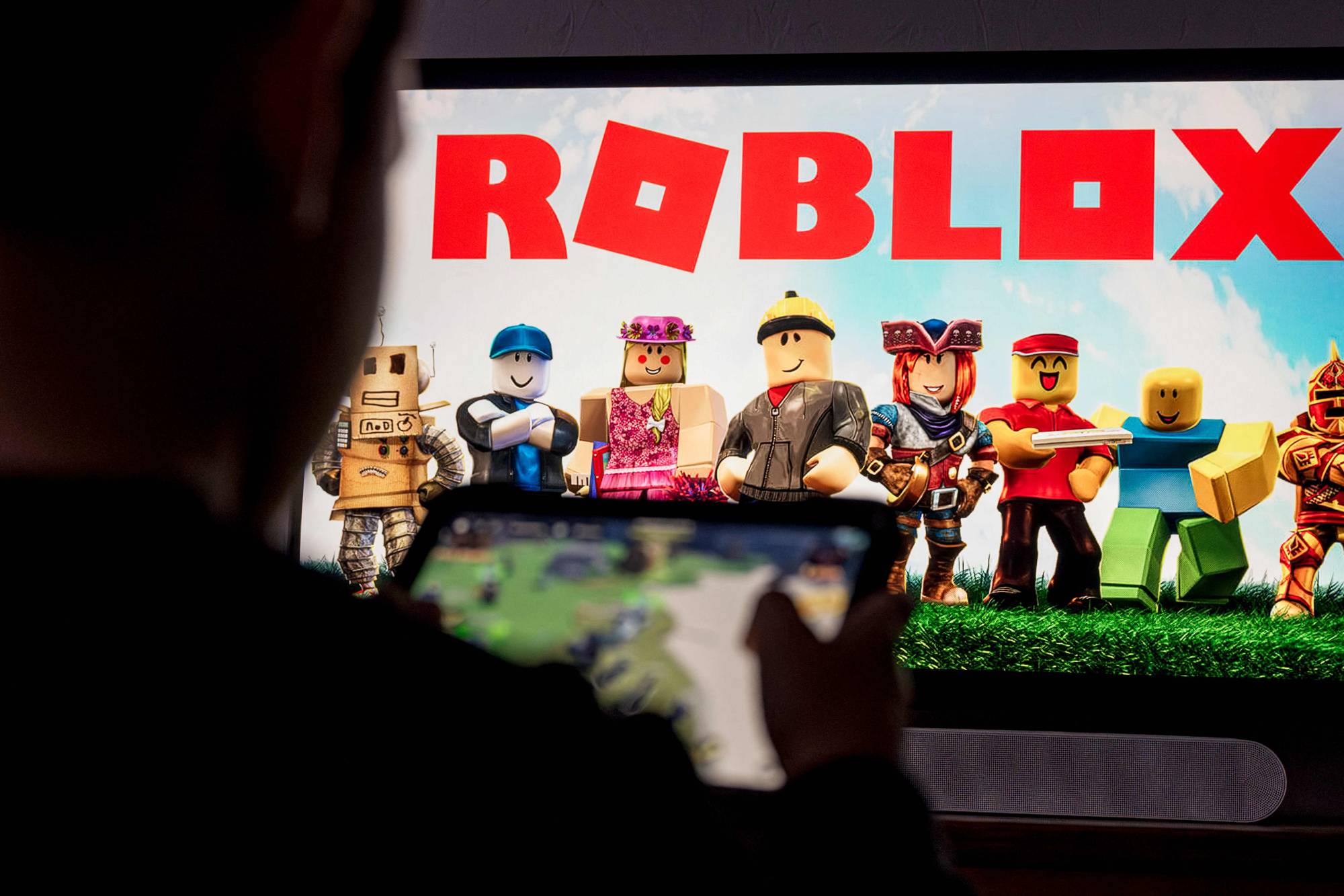 En person holder en nettbrett og spiller. På nettbrettet vises spillfigurene. Bak nettbrettet vises en stor Roblox-logo. (Bildebeskrivelsen er laget av en KI-tjeneste)