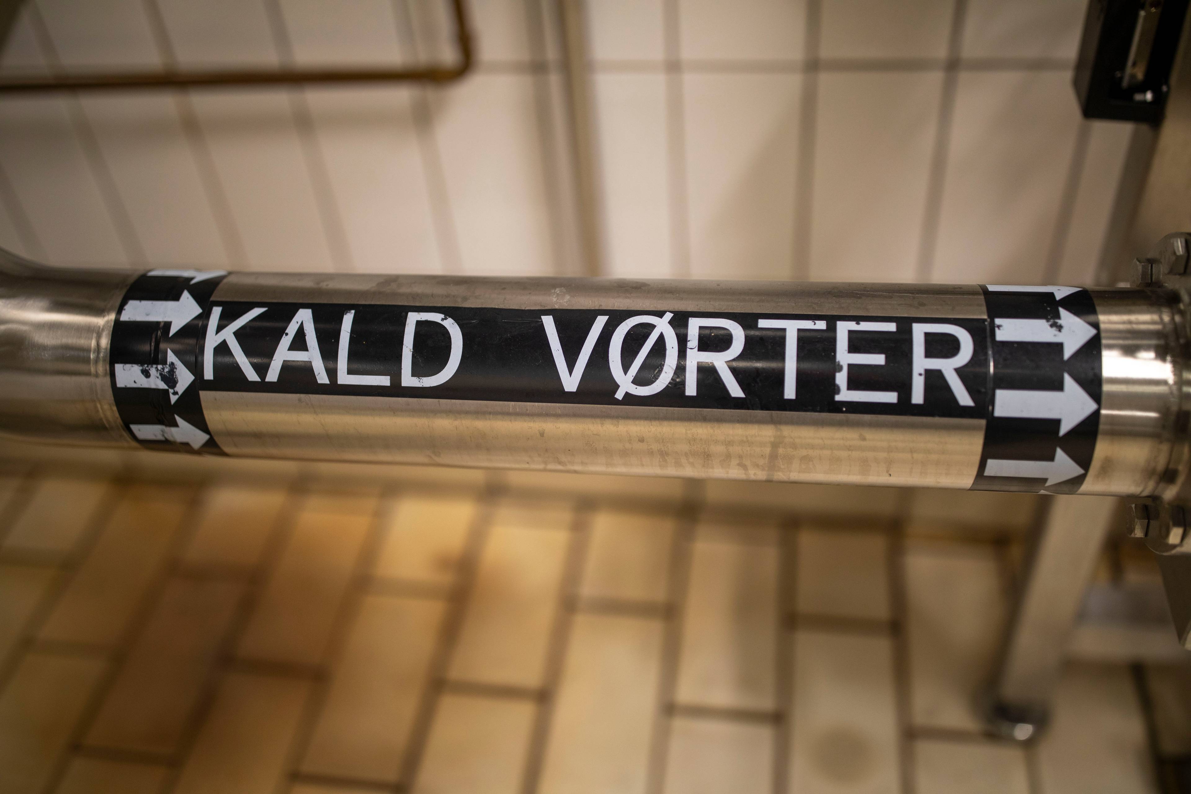 Et rør med teksten "KALDT VØRTER" er synlig. Teksten er på et svart bånd med hvite bokstaver. Røret er laget av rustfritt stål. Bakgrunnen viser en flislagt gulvflate og deler av rør og vegg.