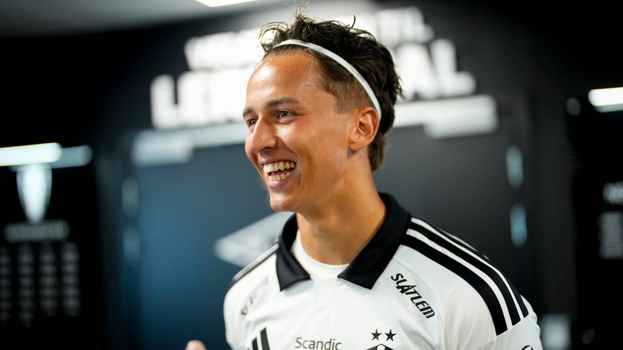 Smilende Rosenborg-spiller i hvit drakt med svart tekst og logoer. (Bildebeskrivelsen er laget av en KI-tjeneste)