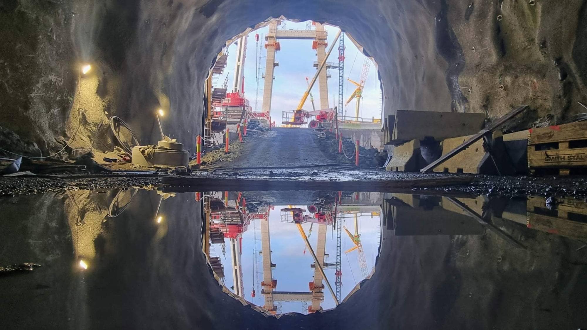 Bildet viser utsikten fra innsiden av en tunnel, med et klart syn på den åpne enden. I det fjerne er det byggemaskiner og kraner synlige, samt en grusvei som fører ut av tunnelen. Det er refleksjoner av de omkringliggende strukturene i en vannpøl på bakken. Omgivelsene er preget av grovt terreng og arbeidselementer som indikerer konstruksjonsarbeid.