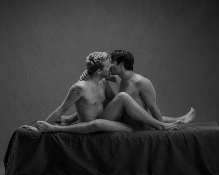 To nakne menn sitter med bena innviklet i hverandre mens de kysser på senga