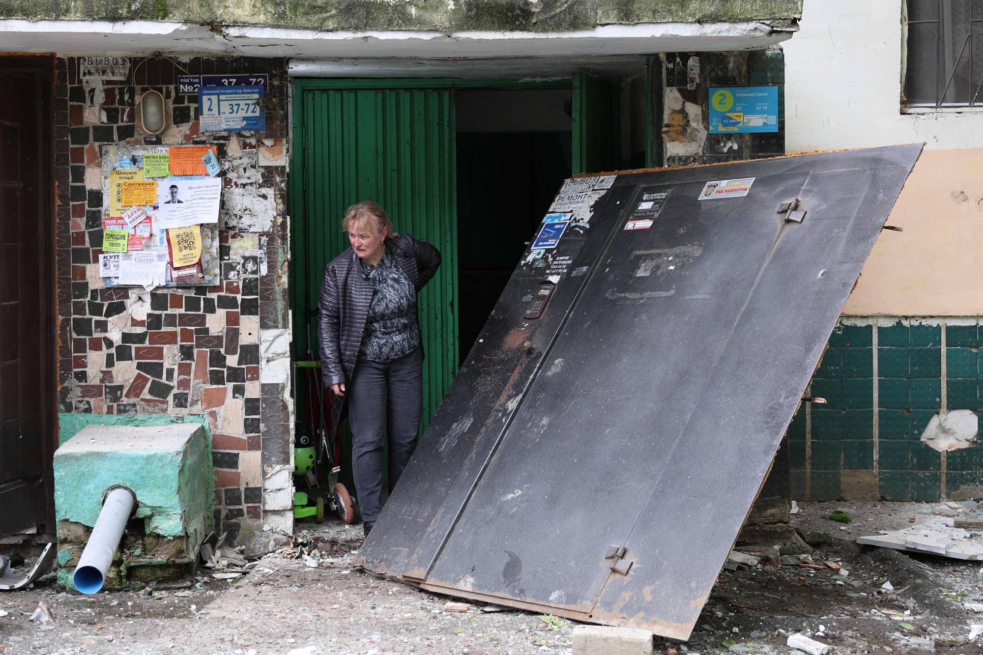 En kvinne står ved en åpen inngang. En stor, sort metalldør ligger skrått ved siden av inngangen. Bakgrunnen viser en bygningsfasade med grønn dør og oppslagstavle. Rundt inngangen er det rusk og grus på bakken. (Bildebeskrivelsen er laget av en KI-tjeneste)
