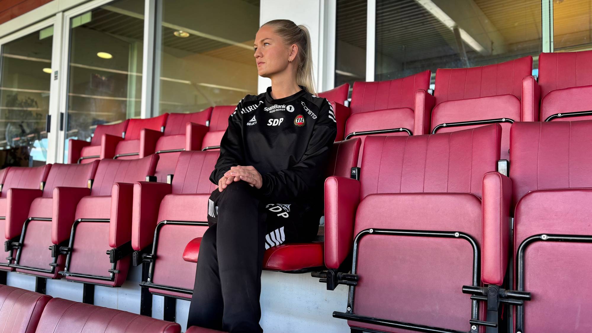 Kvinne i svart treningstøy sitter alene på rader med røde seter på en tribune. (Bildebeskrivelsen er laget av en KI-tjeneste)
