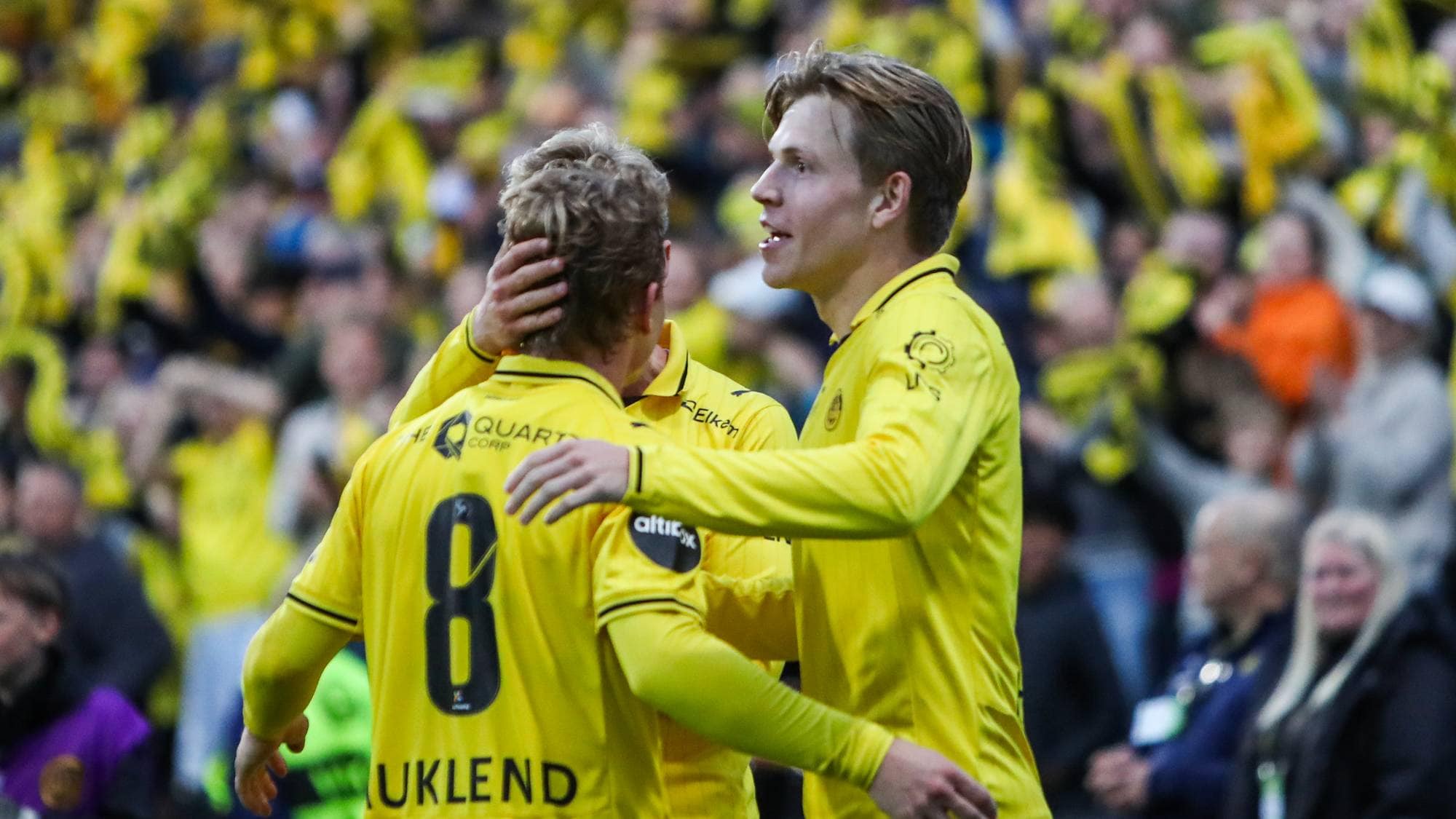 To fotballspillere i gule drakter feirer sammen foran publikum. (Bildebeskrivelsen er laget av en KI-tjeneste)