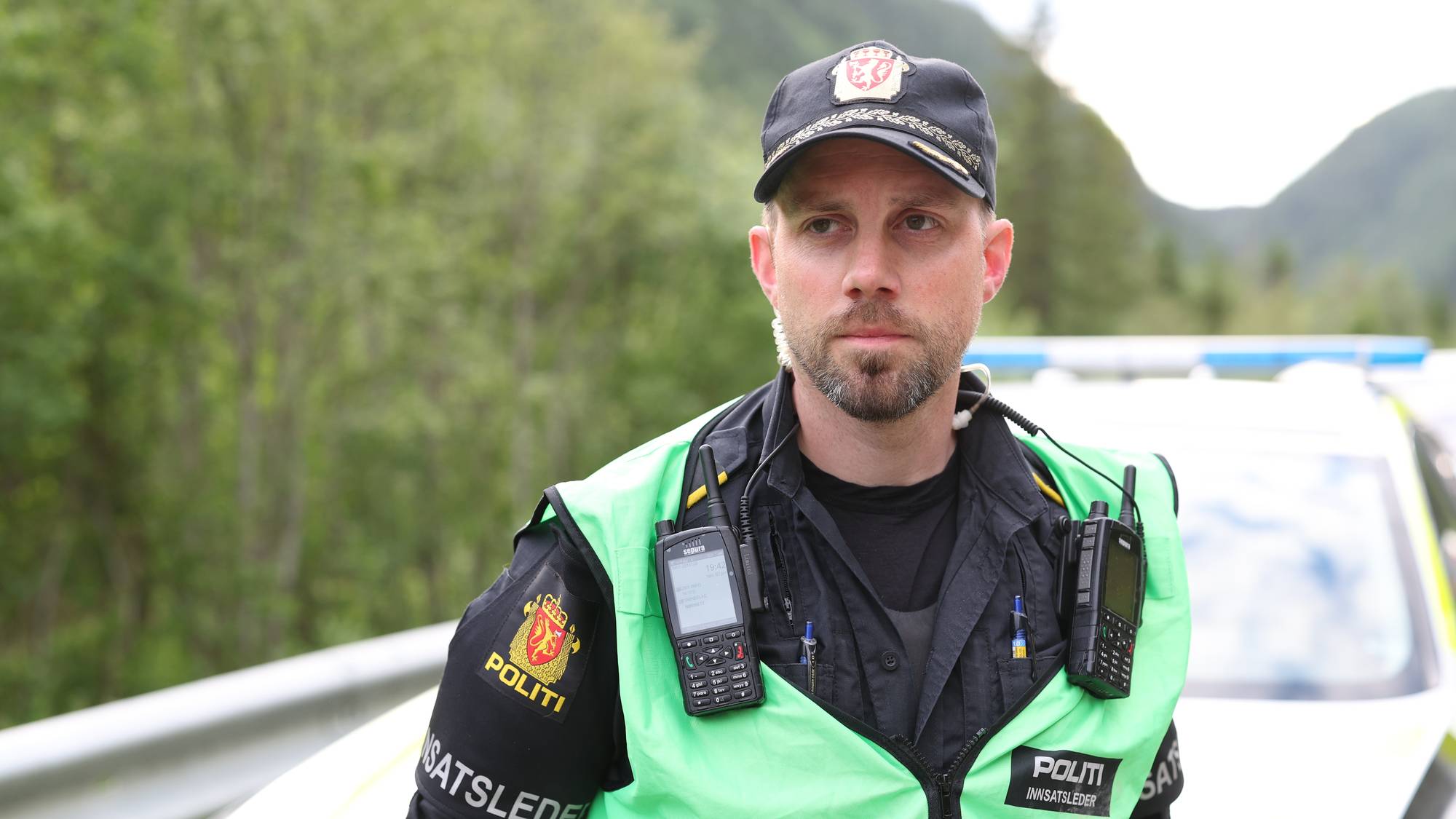 En politimann står ved siden av en politibil. Han har på seg politiuniform med vest. Bakgrunnen er utydelig og viser et grønt skogholt og fjell. Mannen ser til høyre for kameraet. (Bildebeskrivelsen er laget av en KI-tjeneste)