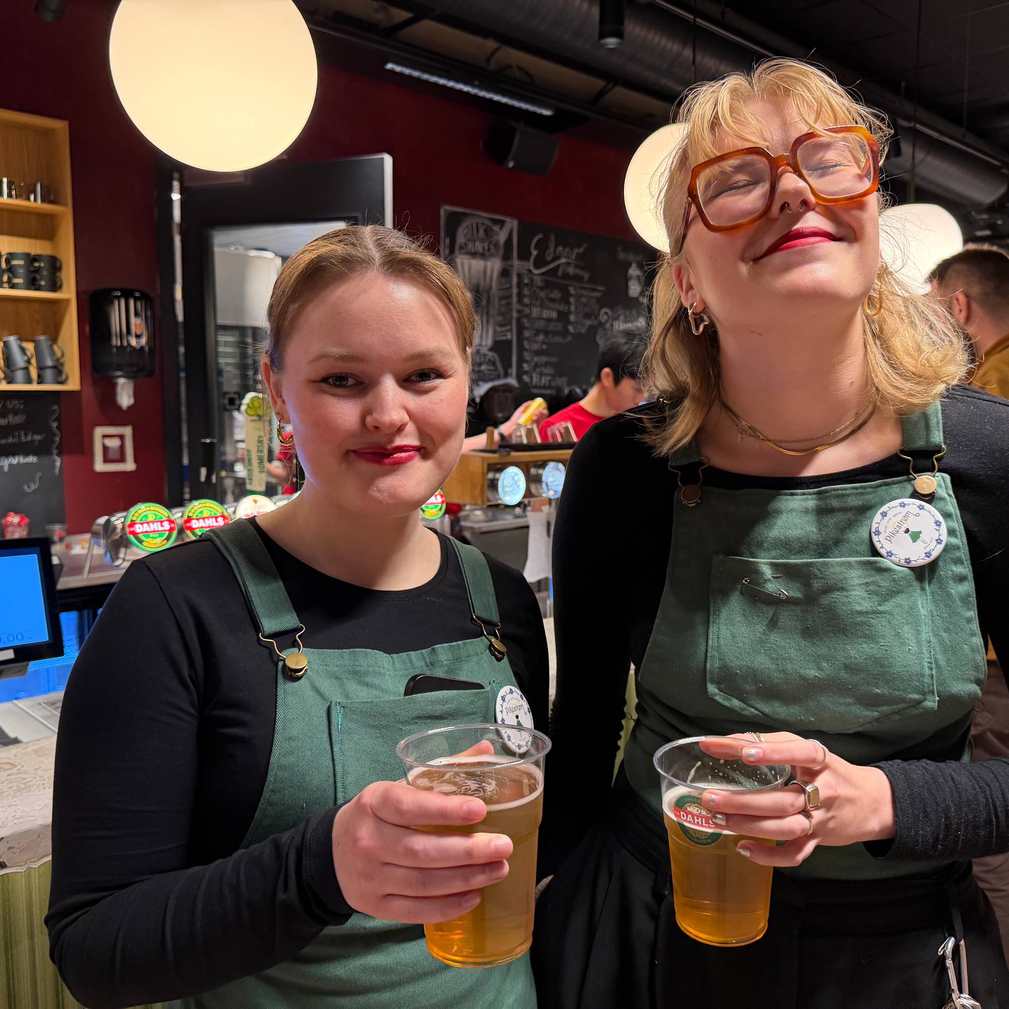 To kvinner i forkle holder glass med øl bak en bar. (Bildebeskrivelsen er laget av en KI-tjeneste)