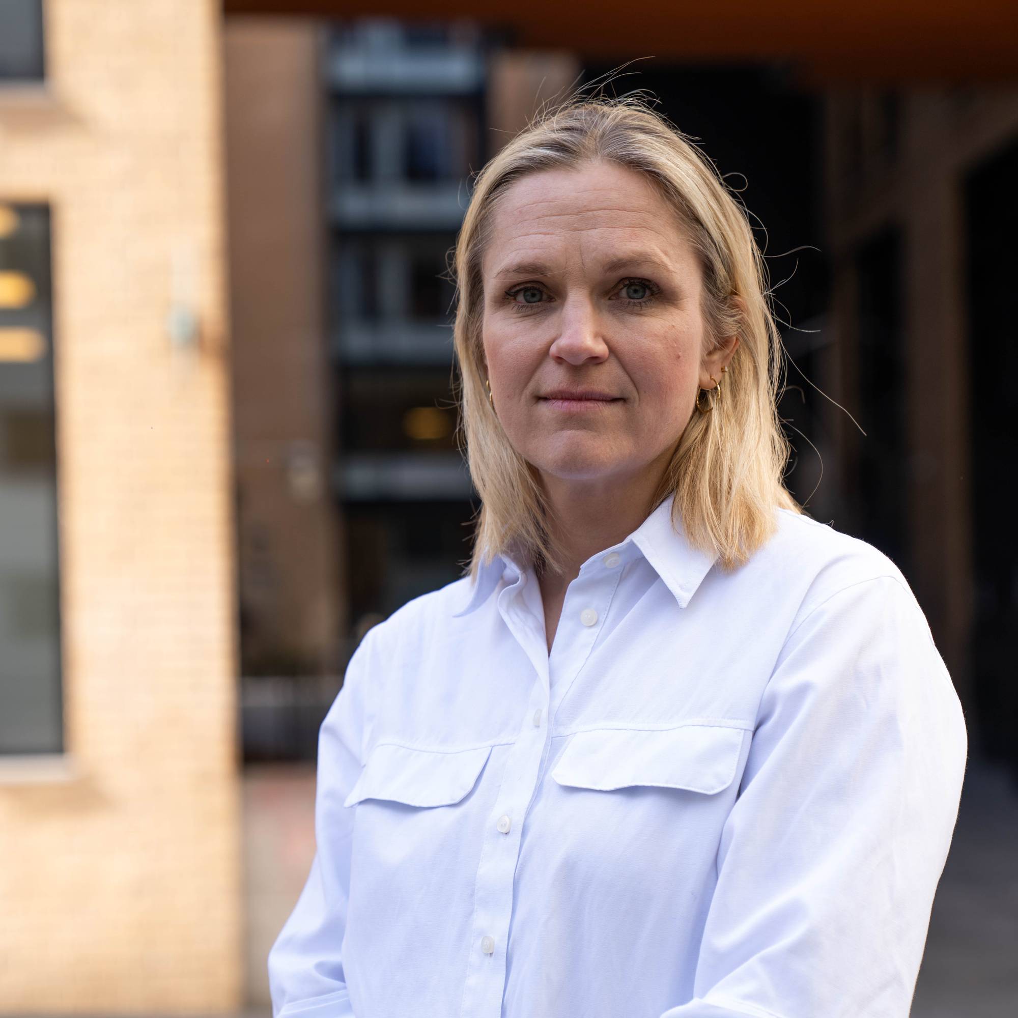 En portrett av blond kvinne i hvit skjorte, svarte bukser som har hendene samlet og kikker i kamera.