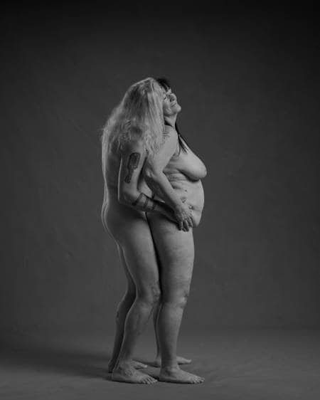 En naken mann med langt hvitt hår står bak en naken kvinne med langt sort hår. Hun lener seg mot ansiktet hans og han holder henne på hoftene. De er et godt voksent par.