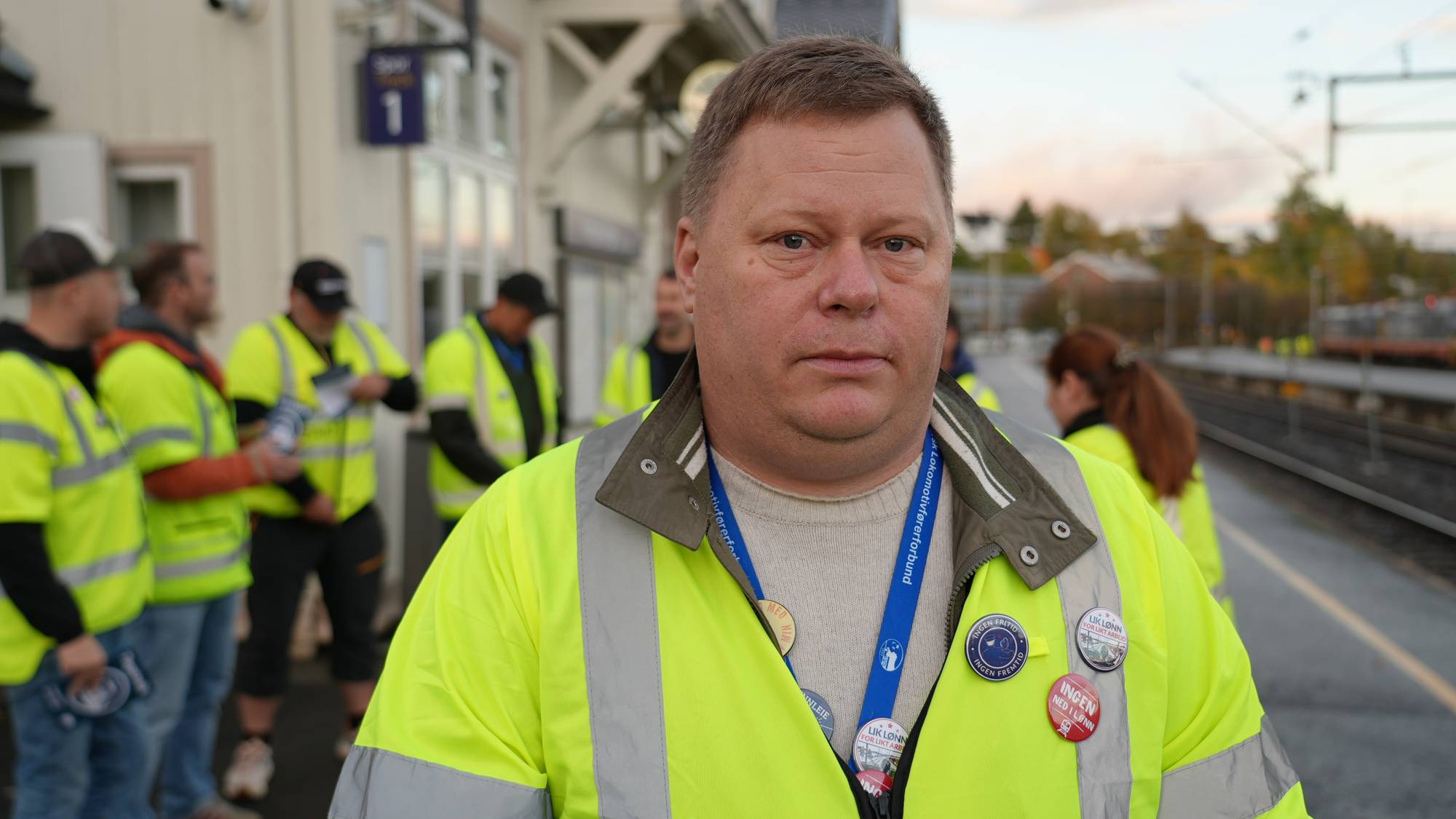 En mann i gul refleksvest står på en togstasjon omgitt av andre personer i lignende bekledning. (Bildebeskrivelsen er laget av en KI-tjeneste)