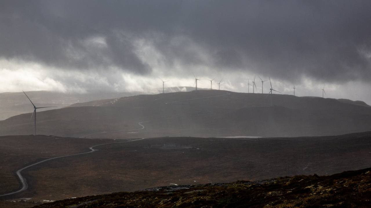 Et ultratotalt utsnitt av Roan vindkraftverk, sett på avstand. I forgrunnen ser man en vei som snor seg gjennom landskapet. I bakgrunnen skimter man vindturbiner som stikker opp i horisonten mellom grå skyer.