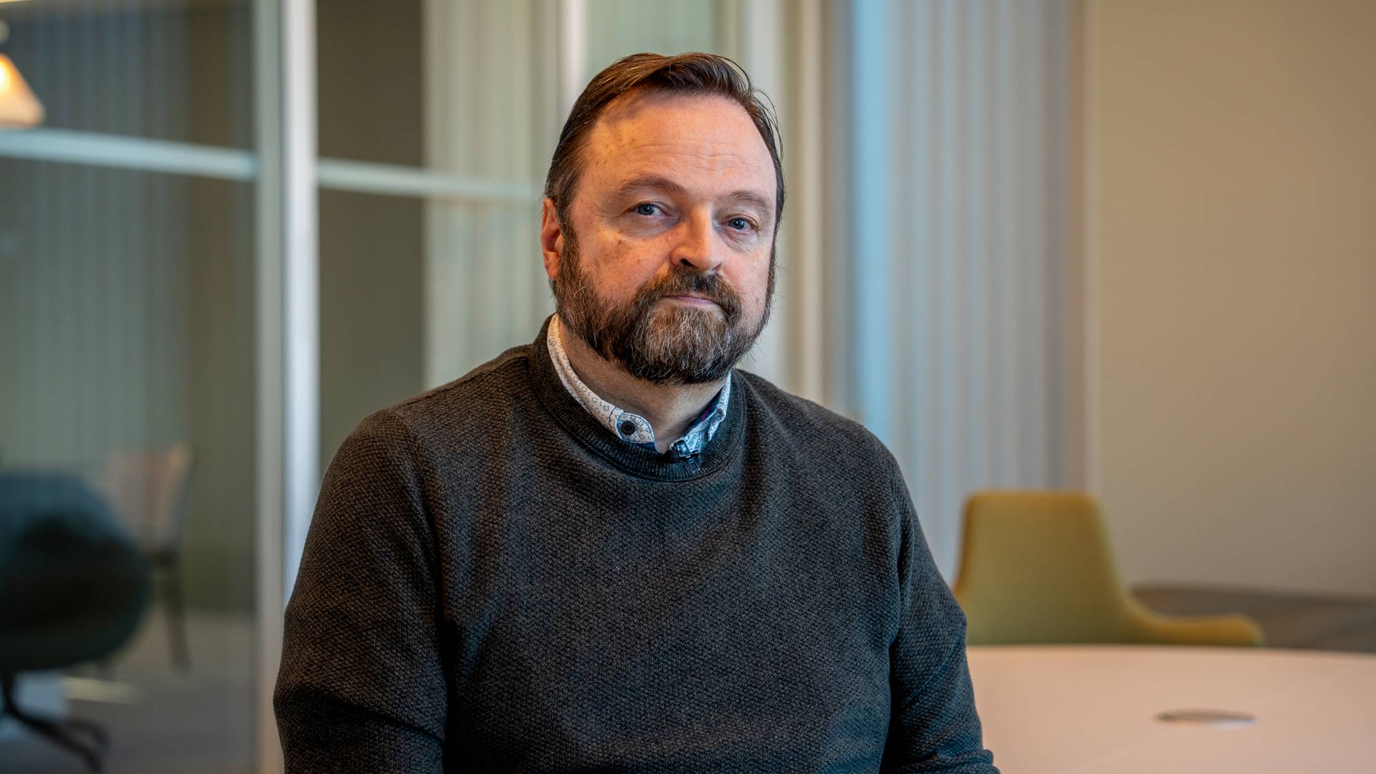 Mann med skjegg og mørk genser sitter i et kontorlandskap. (Bildebeskrivelsen er laget av en KI-tjeneste)