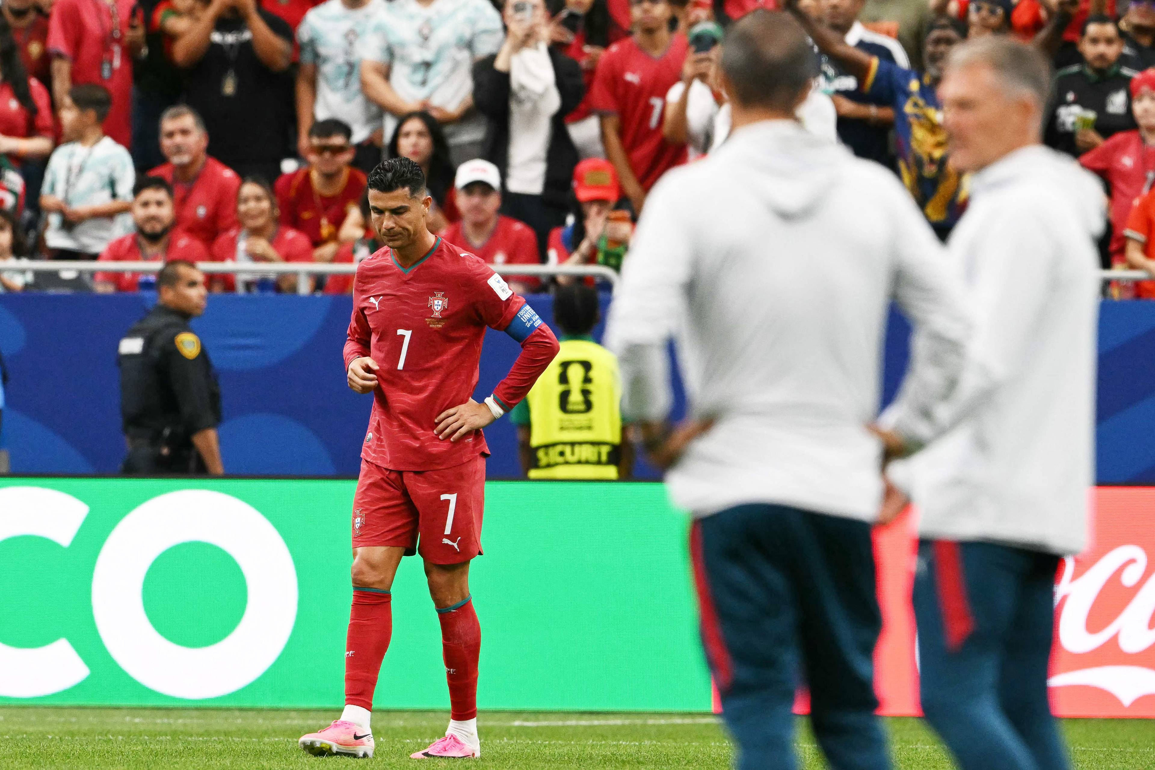 En fotballspiller med draktnummer 7 er på banen med armene i siden. Bak ham står det et sikkerhetspersonell og tilskuere på tribunen. To andre personer, sannsynligvis trenere, går forbi. (Bildebeskrivelsen er laget av en KI-tjeneste)