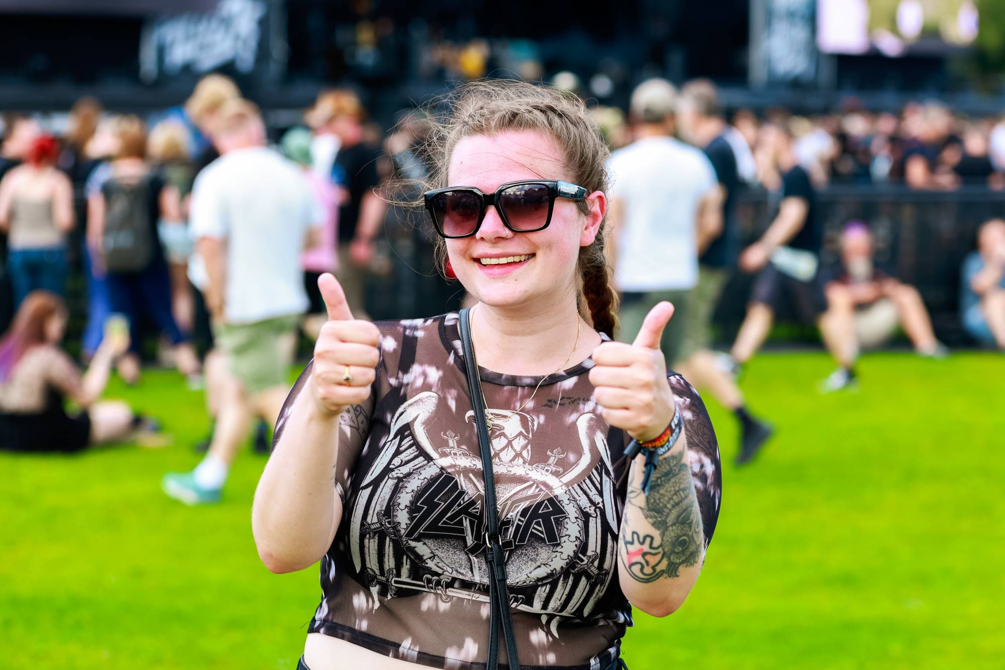 Tone Line gir tommelen opp på festival. Hun har på seg en gjennomsiktig topp med et bandlogo. Bak henne er en folkemengde med andre festivaldeltakere. (Bildebeskrivelsen er laget av en KI-tjeneste)
