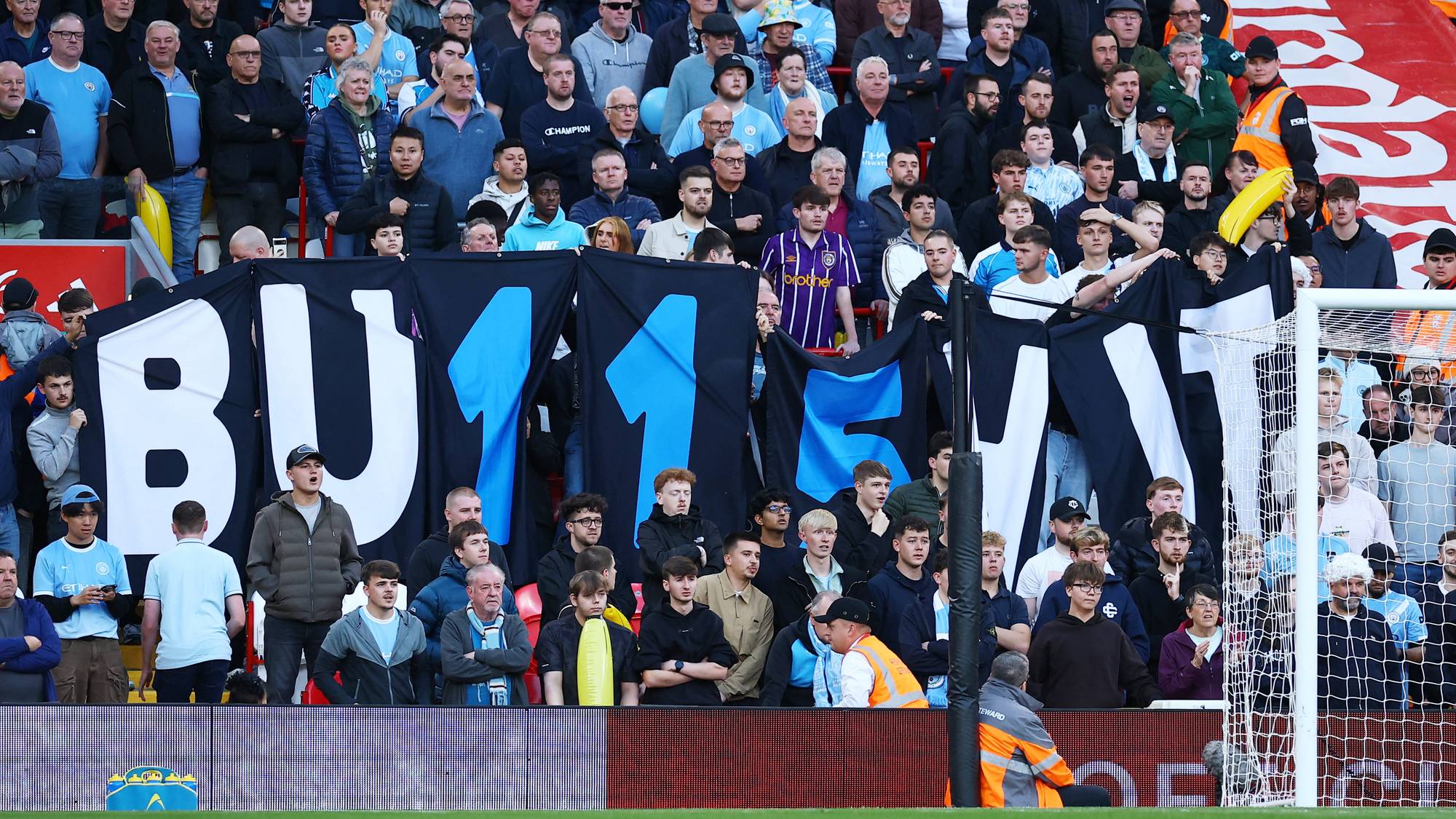 Publikum på en fotballtribune holder opp et stort svart banner med påskriften BU11M1T. (Bildebeskrivelsen er laget av en KI-tjeneste)