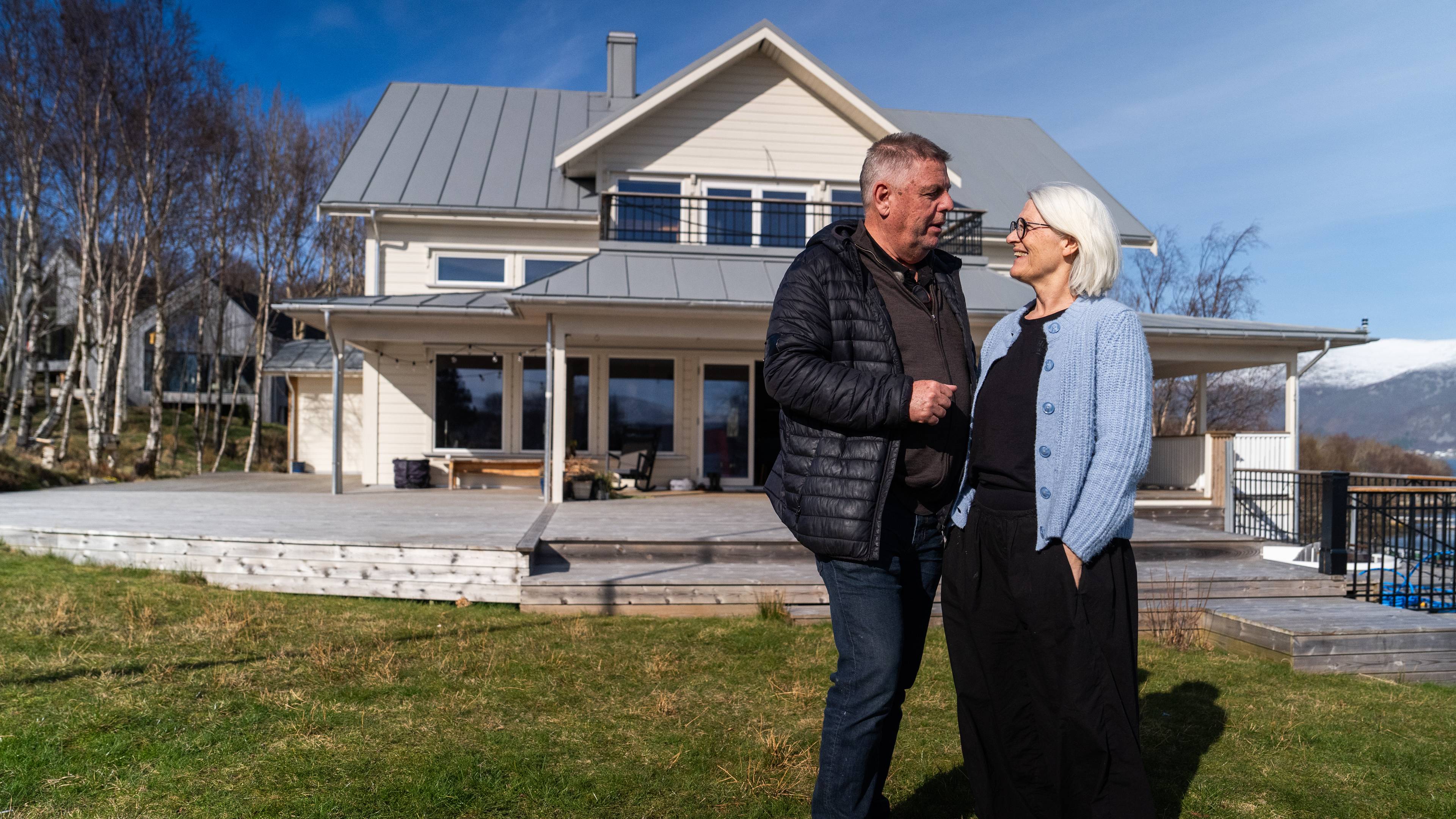 Et eldre ektepar står foran et stort hvitt hus med store vinduer. Paret ser på hverandre og smiler. Bak dem er en stor terrasse og et grønt gressfelt. Huset er to etasjer høyt og har et grått saltak. Ekteparet er kledd i uformelle klær. (Bildebeskrivelsen er laget av en KI-tjeneste)