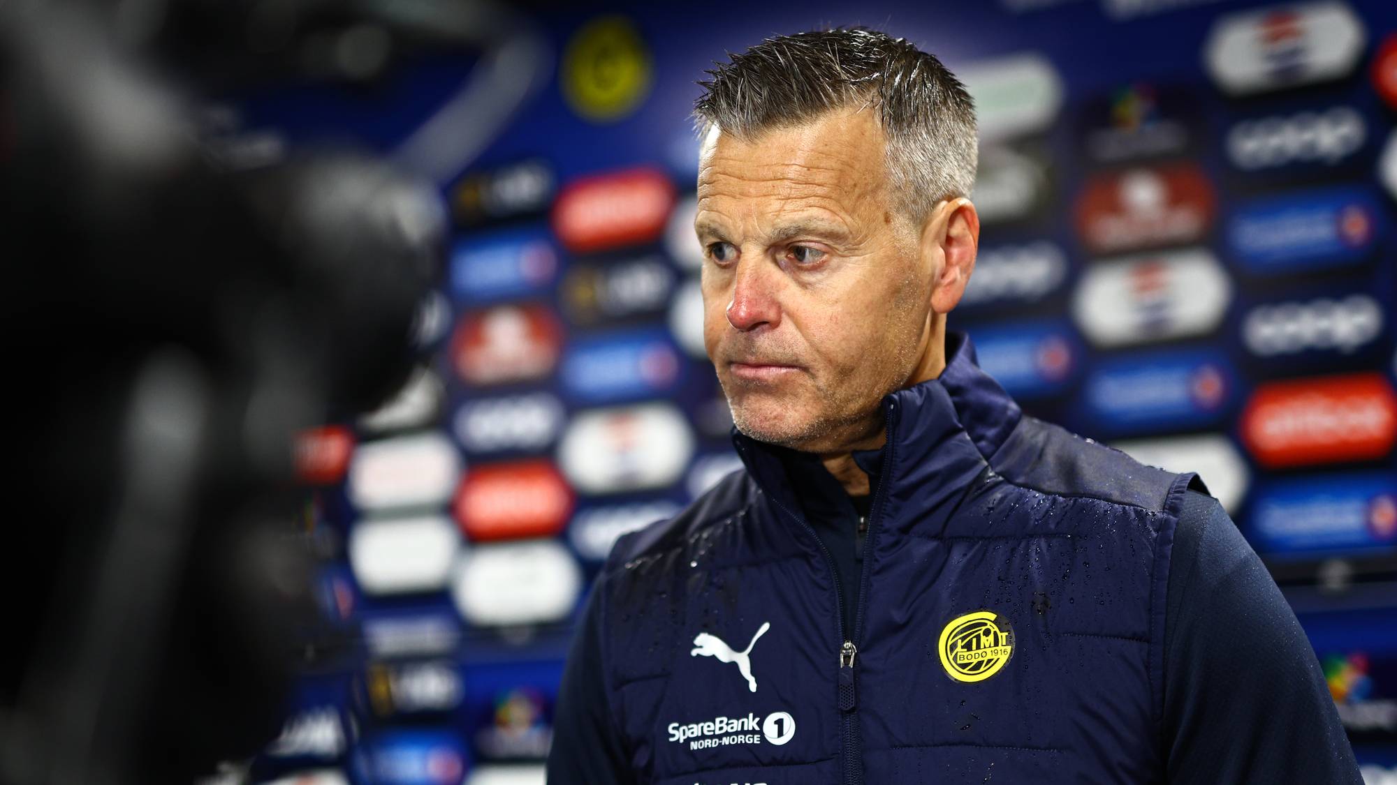 En mann i mørk vest med logoer ser til siden foran en bakvegg med sponsoravtaler. Mannen har grått hår og ser alvorlig ut. Bakgrunnen er et sett med forskjellige sponsorlogoer på en blå vegg. Mannen er antagelig en trener eller leder i en idrettsorganisasjon. (Bildebeskrivelsen er laget av en KI-tjeneste)