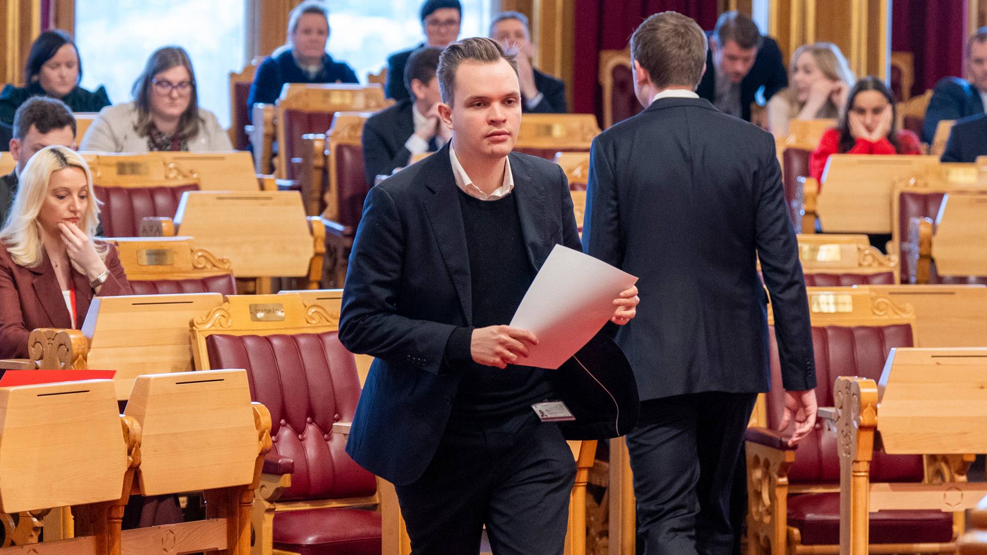 En mann går med papirer i hånden i et stort rom med mange tomme seter. En annen mann går bort fra kameraet. Kvinner sitter på radene bak. (Bildebeskrivelsen er laget av en KI-tjeneste)