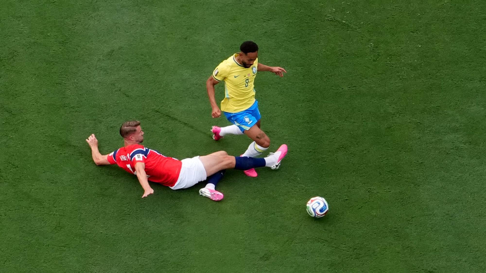 Fotballspiller i gul drakt utfordres av norsk spiller i en takling på gressbanen. (Bildebeskrivelsen er laget av en KI-tjeneste)