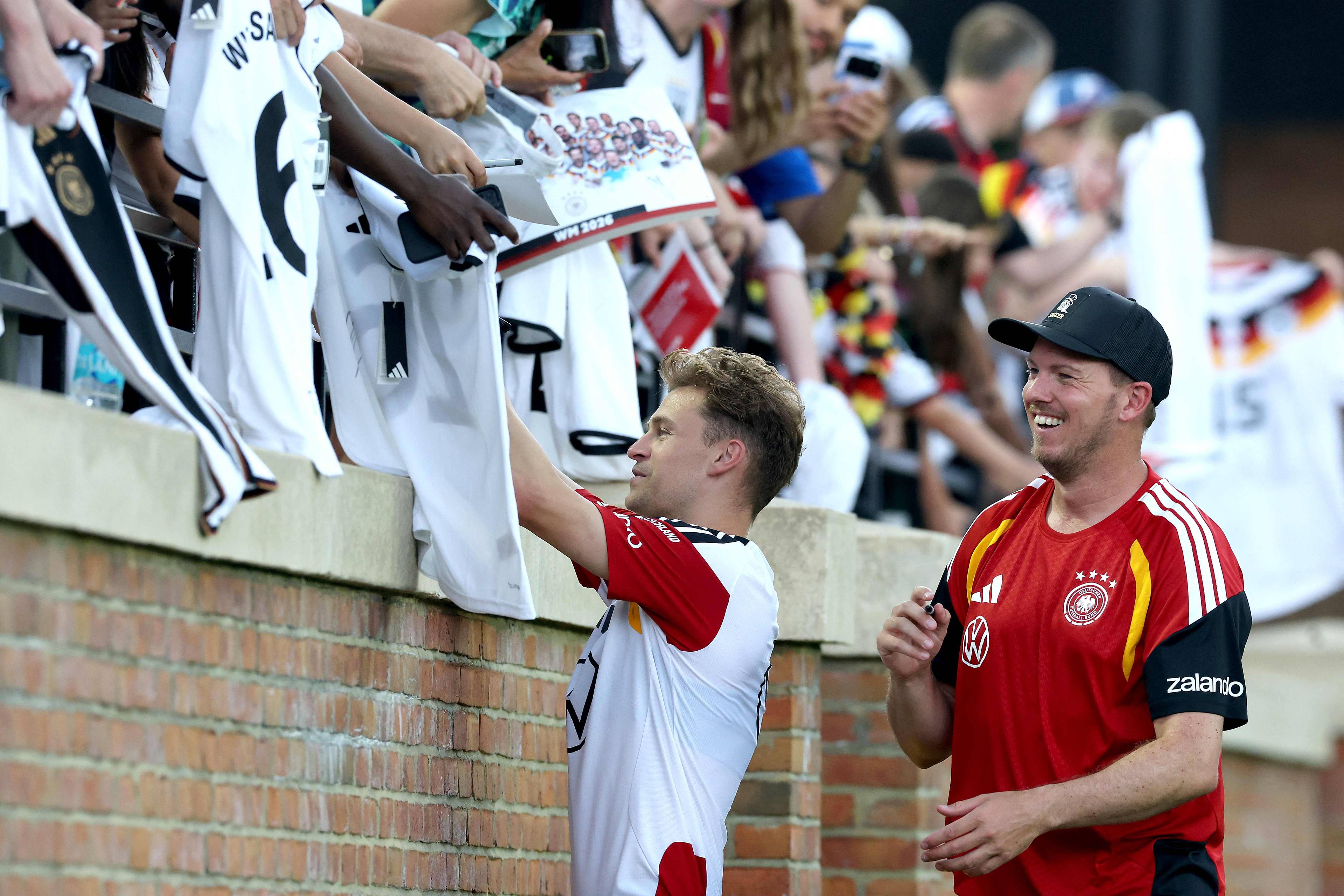 En fotballspiller gir autografer til supportere. En annen mann står ved siden av og smiler. Supporterne holder opp trøyer og autografskilt. Bildet er tatt utendørs foran en murvegg og tribunene. (Bildebeskrivelsen er laget av en KI-tjeneste)