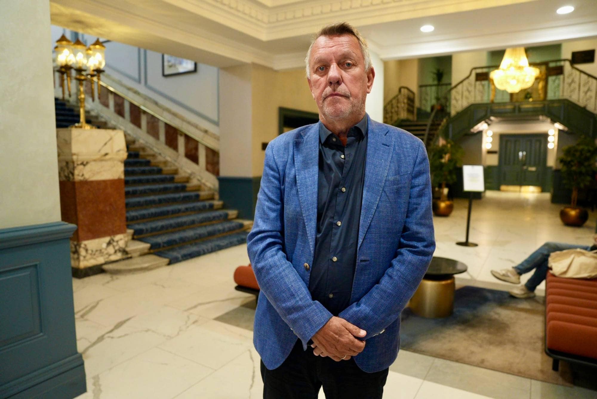 Eldre mann i blå blazer står i en hotellobby. I bakgrunnen ses en trapp med blått teppe. (Bildebeskrivelsen er laget av en KI-tjeneste)
