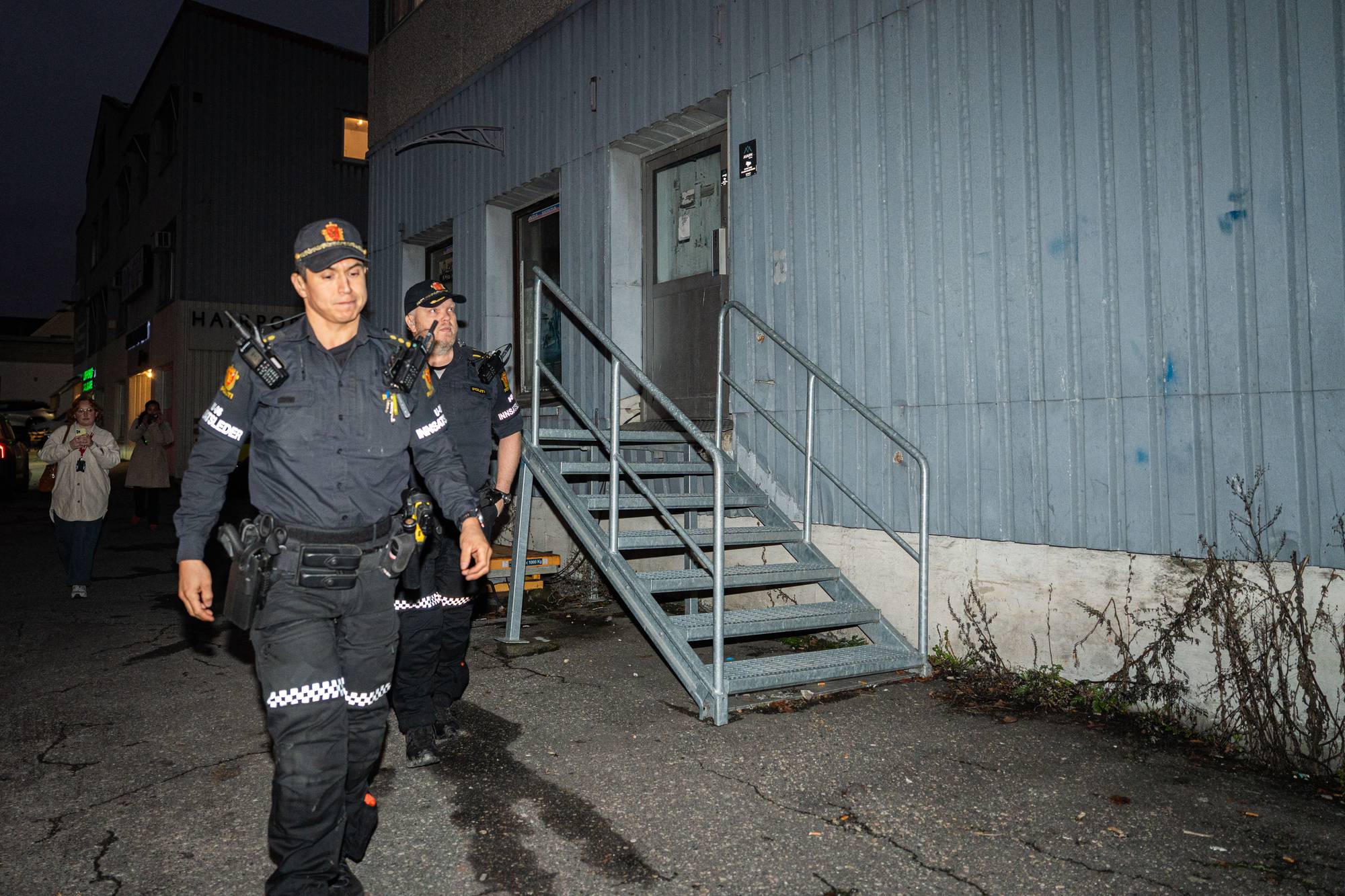 To politibetjenter går i en mørk bakgård nær en bygning med blå vegger. Den ene betjenten er iført sort uniform og utstyr, og de beveger seg mot en trapp. I bakgrunnen er en forbipasserende person med lys jakke. Omgivelsene er preget av nattemørke og urbane elementer.
