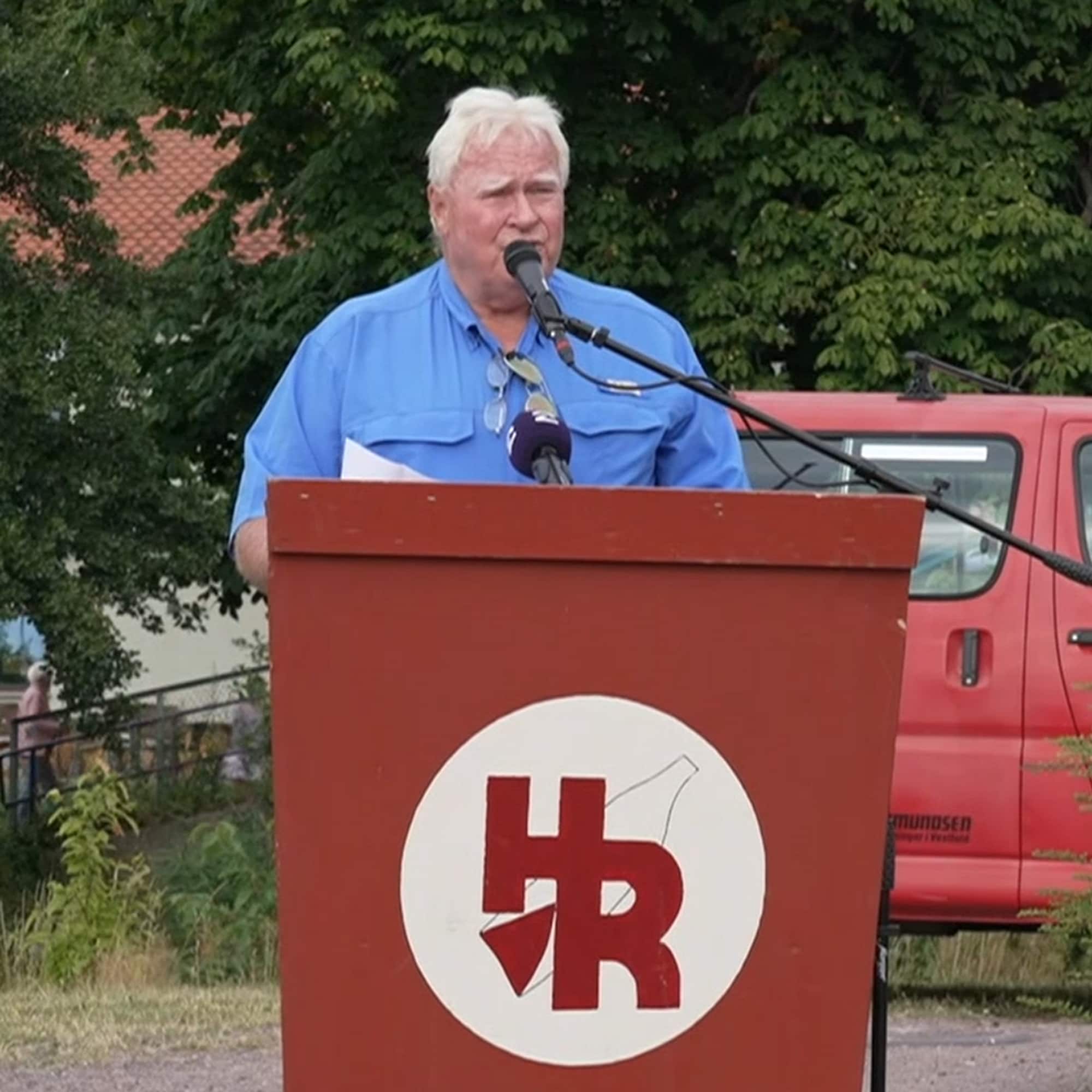 Eldre mann med lyst hår snakker i mikrofon bak en talerstol med HR-logo utendørs.