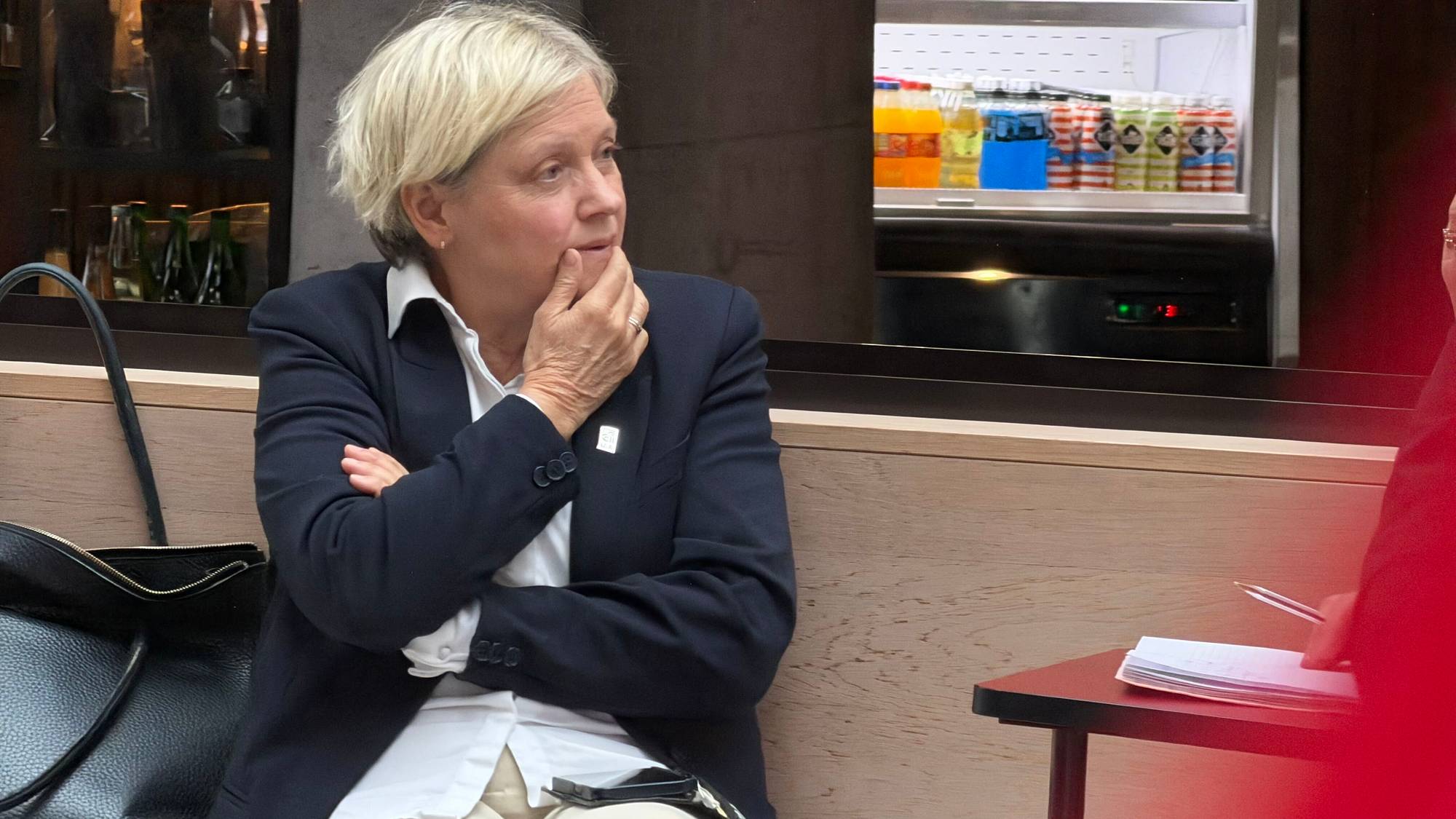 En kvinne med blondt hår, hvit skjorte, blå blazer og beige bukser sitter ved et bord. Hun ser tankefull ut. 