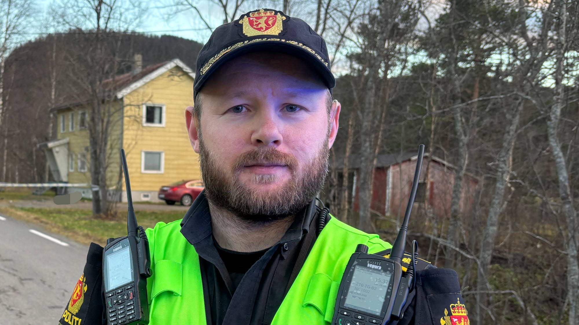 Politibetjent i uniform og gul refleksvest står ved en vei med skog og bebyggelse i bakgrunnen. (Bildebeskrivelsen er laget av en KI-tjeneste)