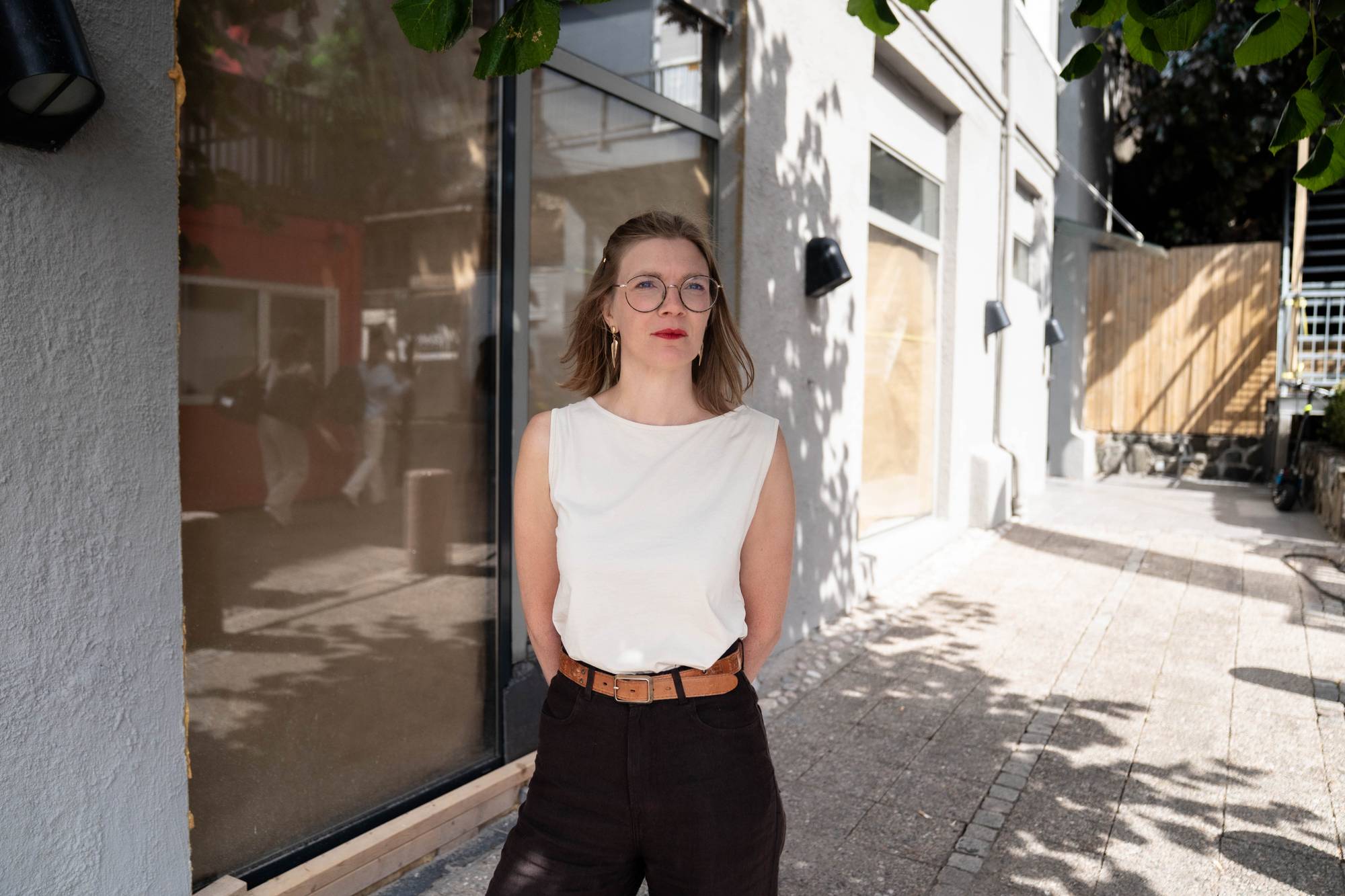 Kvinne med briller og lys topp står utendørs foran en bygning med store vinduer. (Bildebeskrivelsen er laget av en KI-tjeneste)