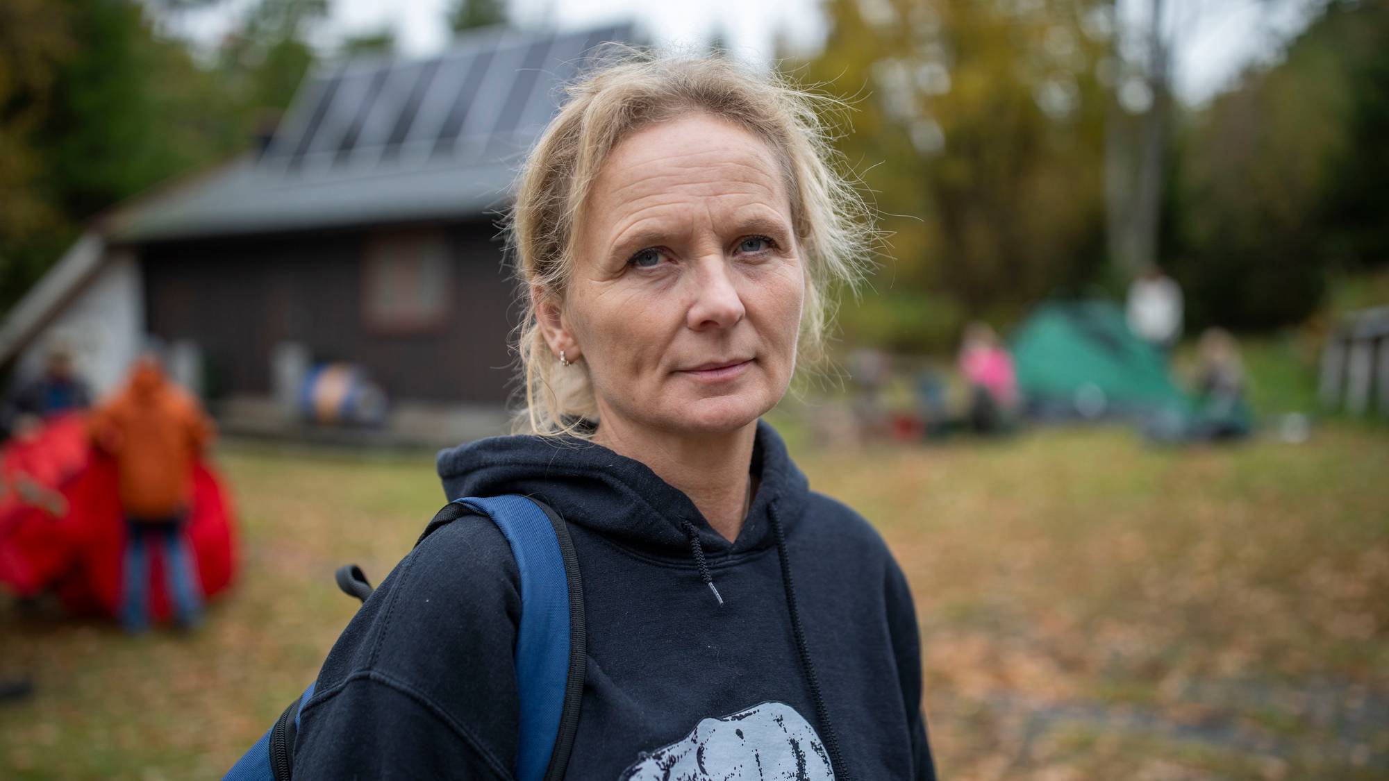 Kvinne i svart hettegenser med bjørnemotiv foran en hytte og telt i skogen. (Bildebeskrivelsen er laget av en KI-tjeneste)