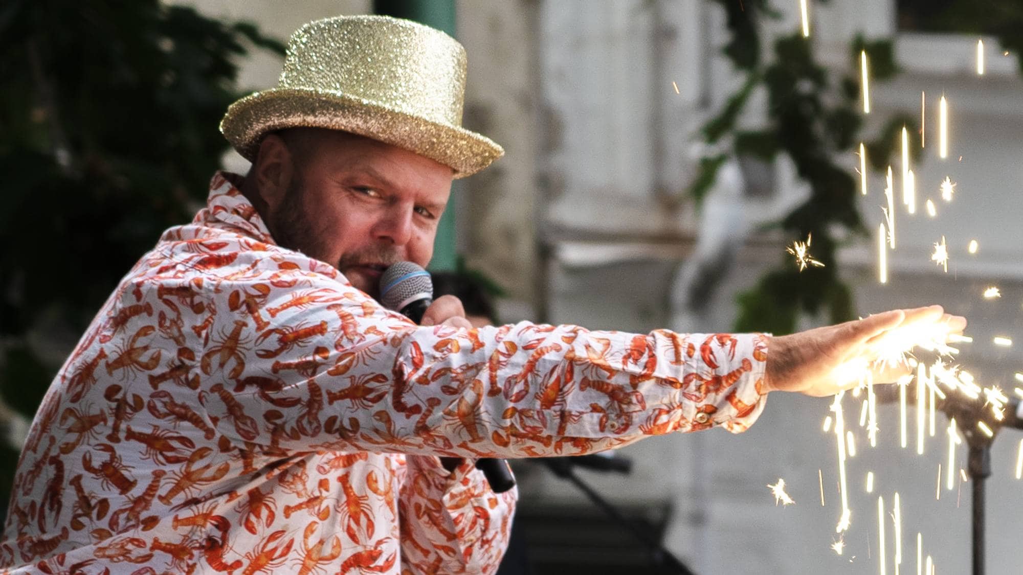 Mann med glitterhatt og hummer-skjorte holder en mikrofon mens han holder frem en gnistrende effekt. (Bildebeskrivelsen er laget av en KI-tjeneste)