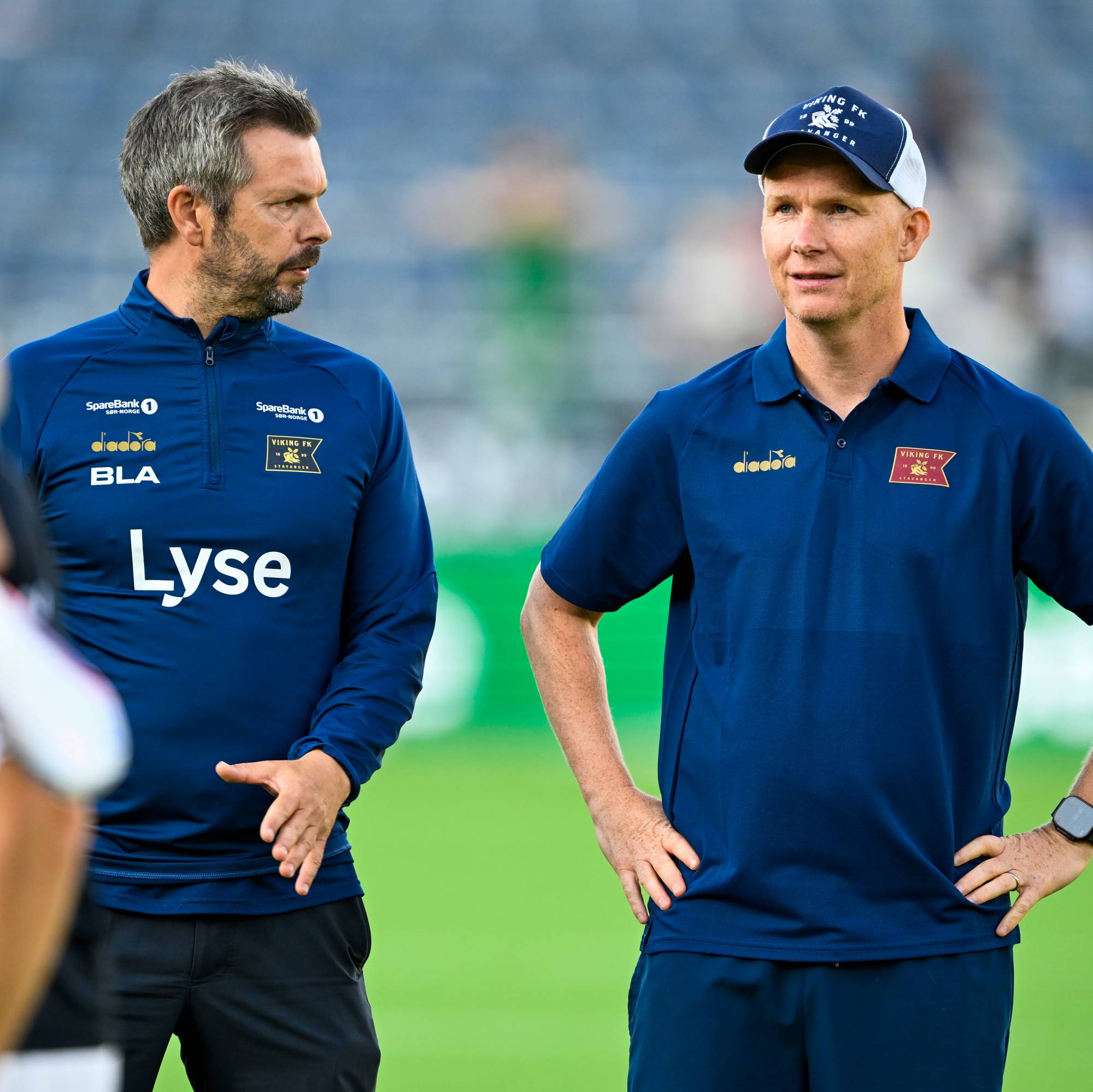 To Viking FK-trenere i mørkeblå bekledning står på en fotballbane. (Bildebeskrivelsen er laget av en KI-tjeneste)