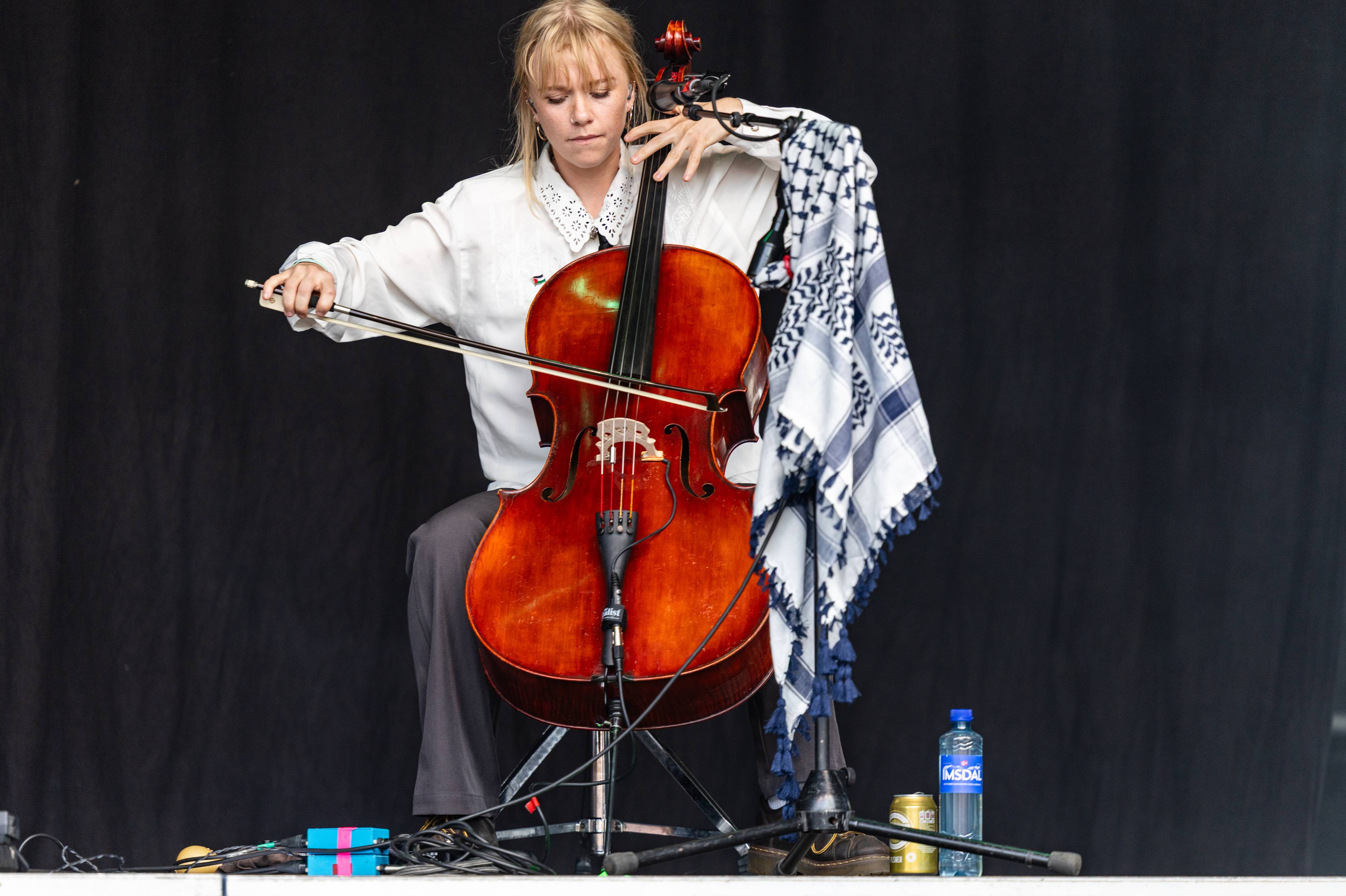Kvinne spiller cello på en scene foran et svart bakteppe. Hun har et palestinaskjerf på instrumentet.