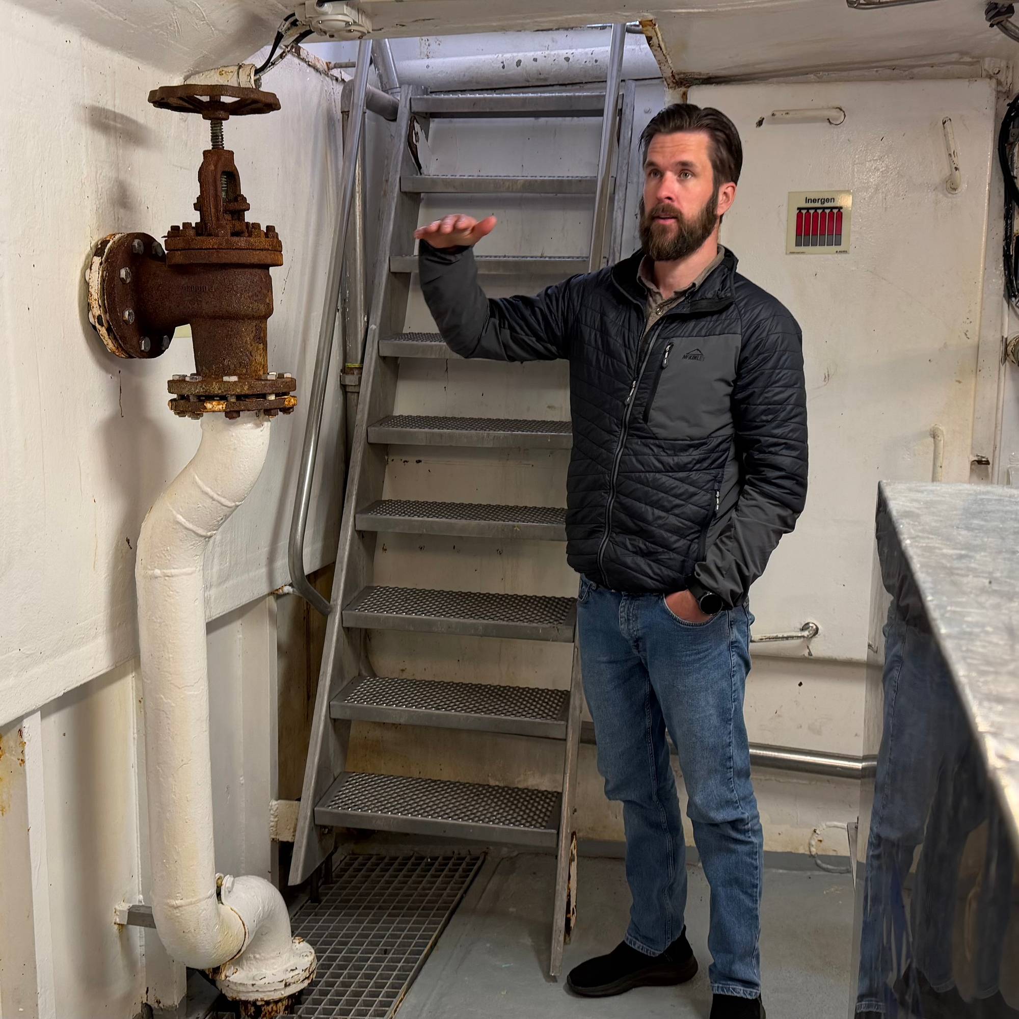 Mann i svart jakke viser vannstand med hånda. Lensepumpa og lensebrønn ved beina. 