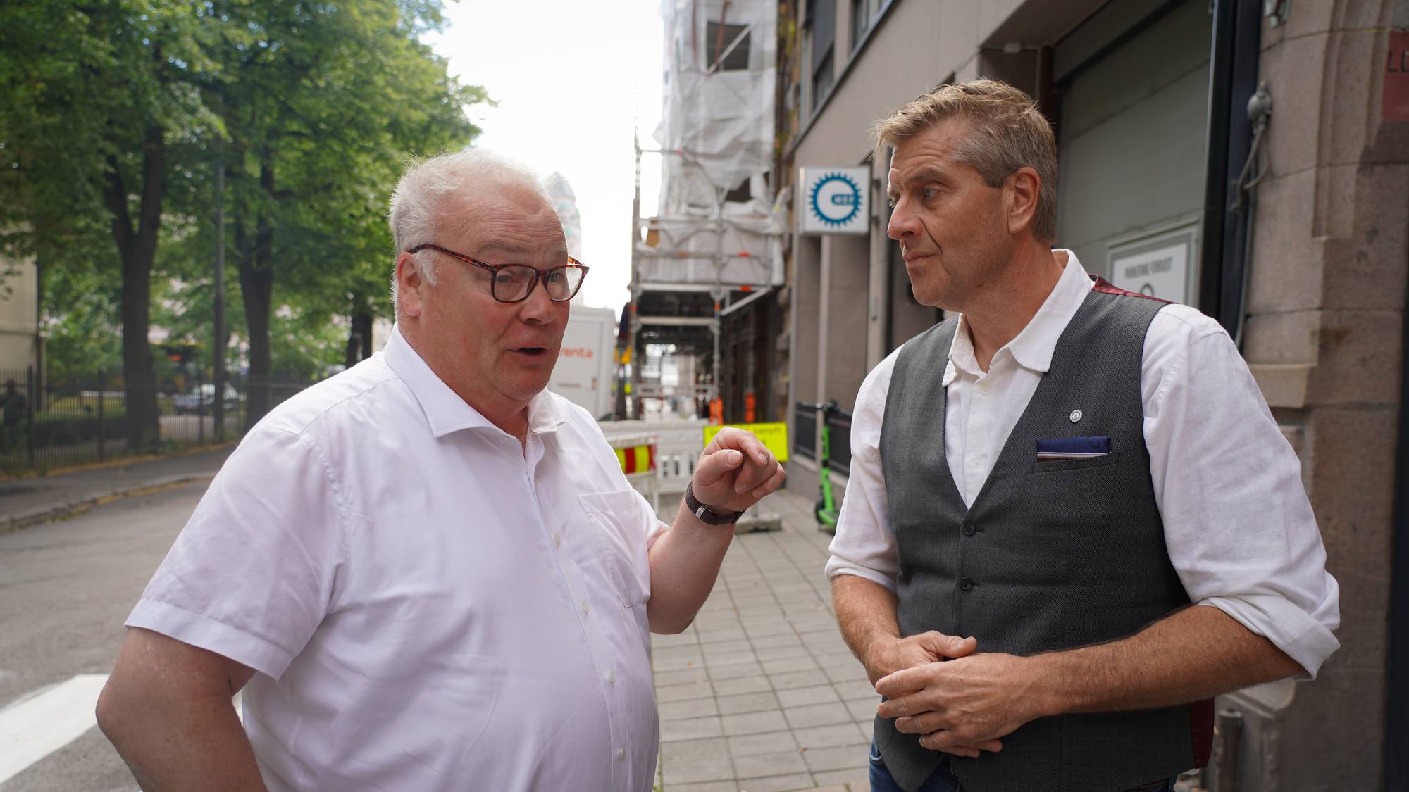 To menn i samtale på en gate. Den ene peker mens den andre lytter i en bygate med byggearbeid. 
