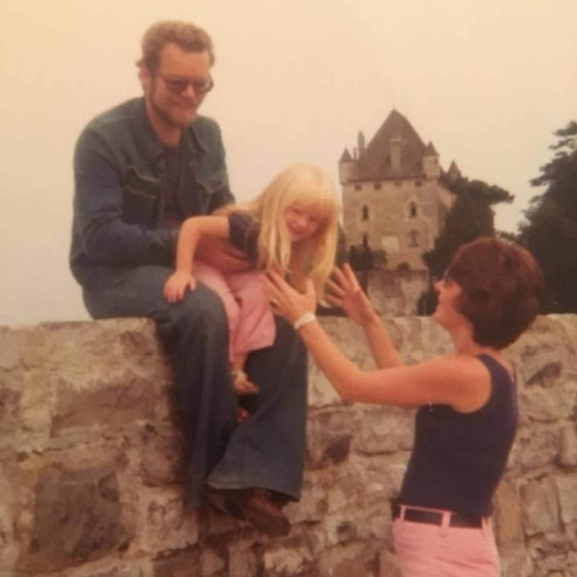 Et bilde tatt på 1970-tallet. En mann sitter på en høy steinmur, han har et barn med lyst hår på fanget, men en kvinne strekker armene ut for å ta henne ned. I bakgrunnen ses et gammelt slottstårn.