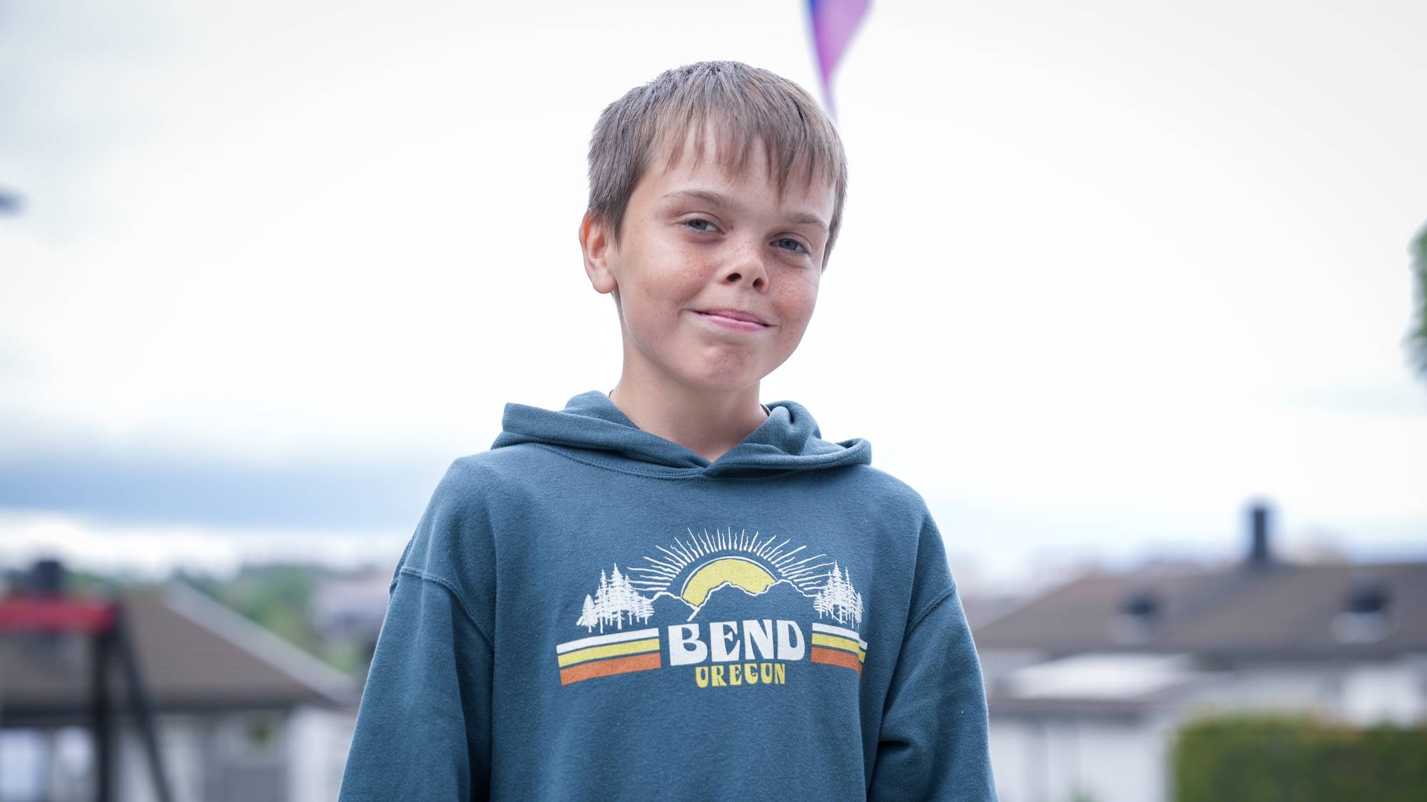 12 år gammel gutt står i skolegård i grønn hettegenser, smiler og ser i kamera. Bak ham ser man tuppen av et regnbueflagg som vaier over ham.