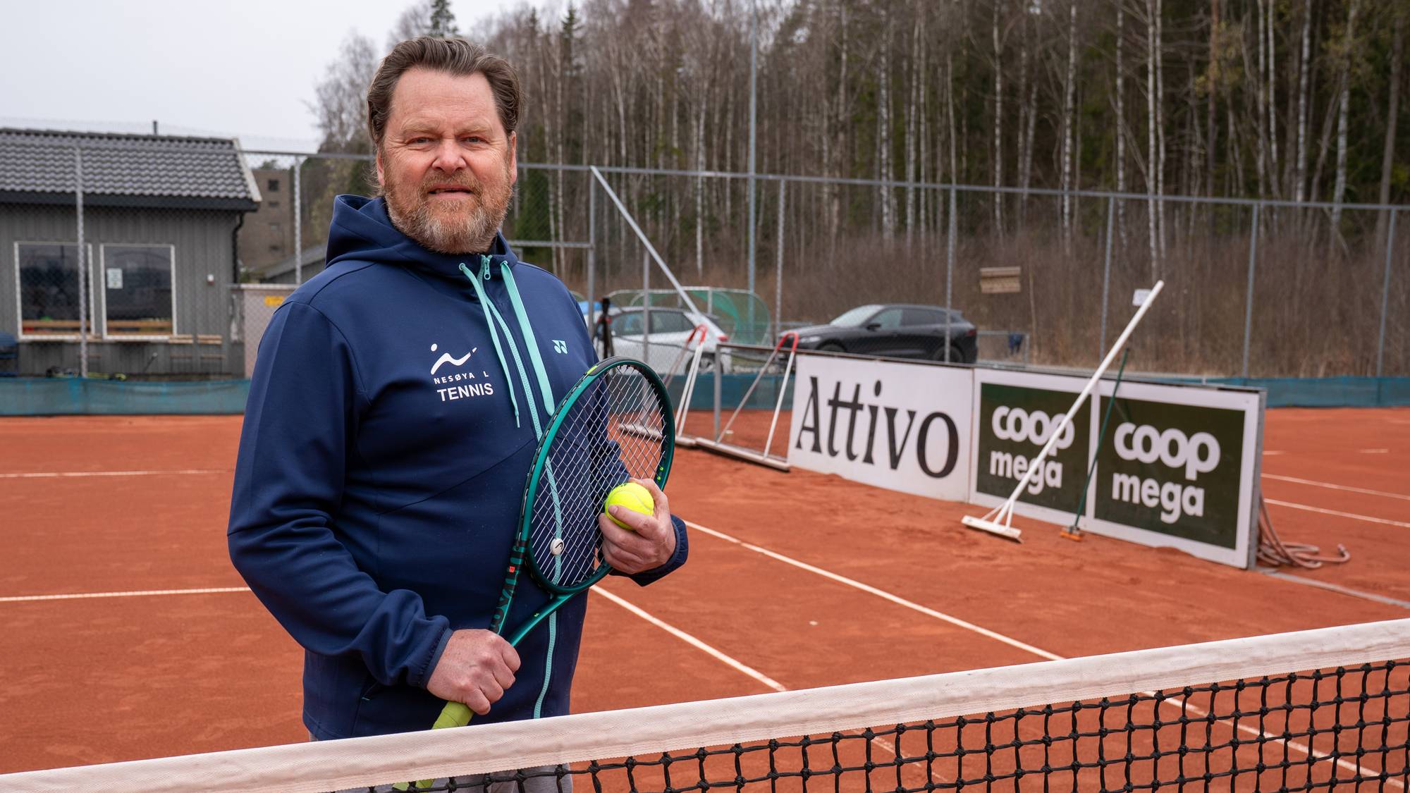 En mann står på en tennisbane med en racket og en ball. Han er kledd i en blå genser og grå bukse. Bak ham er et nett, en bygning, trær og reklameskilt. (Bildebeskrivelsen er laget av en KI-tjeneste)