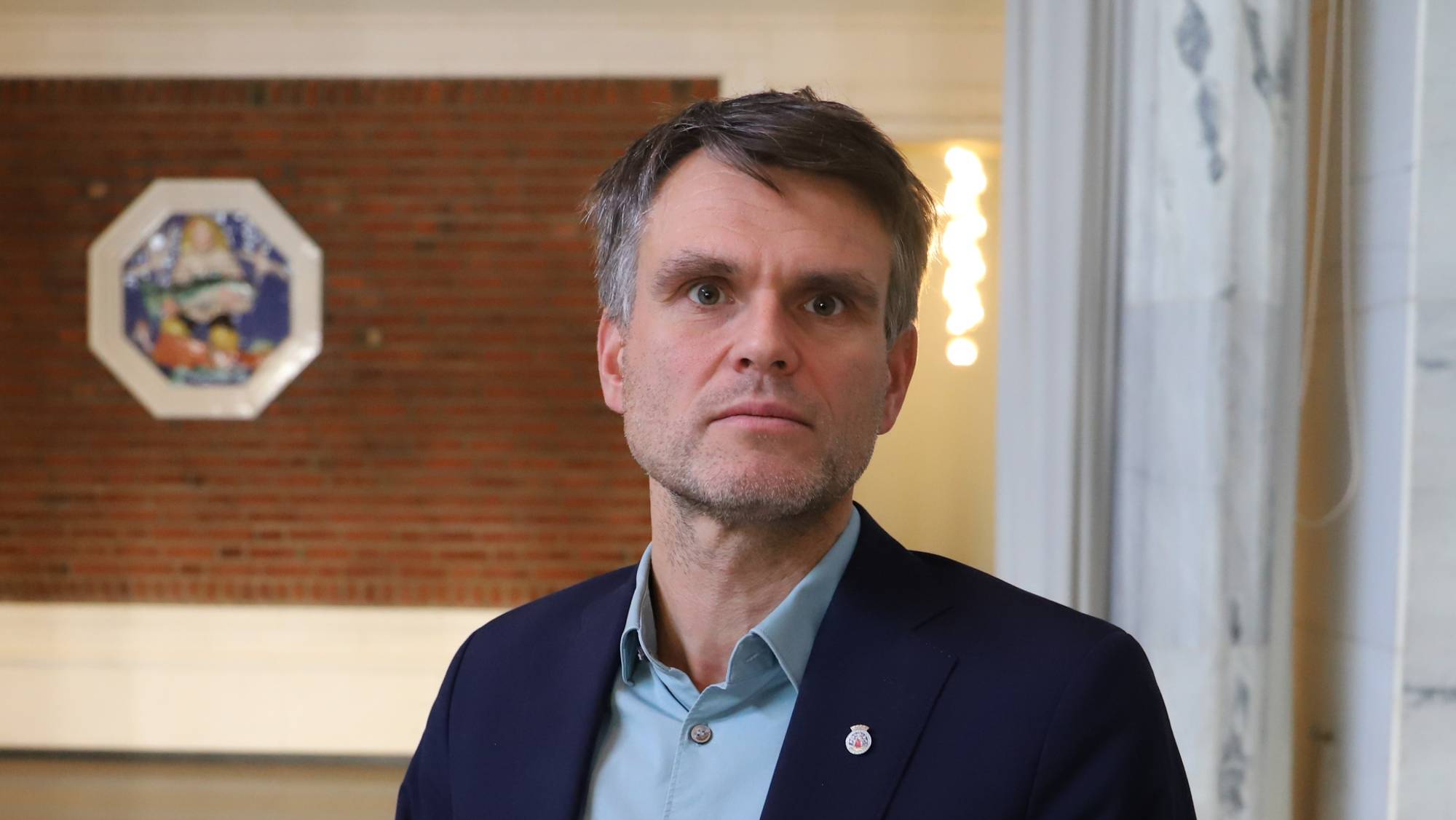 En mann med kort mørkt hår står og ser i kamera, han har på seg en mørk blå dressjakke over en lys blå skjorte.