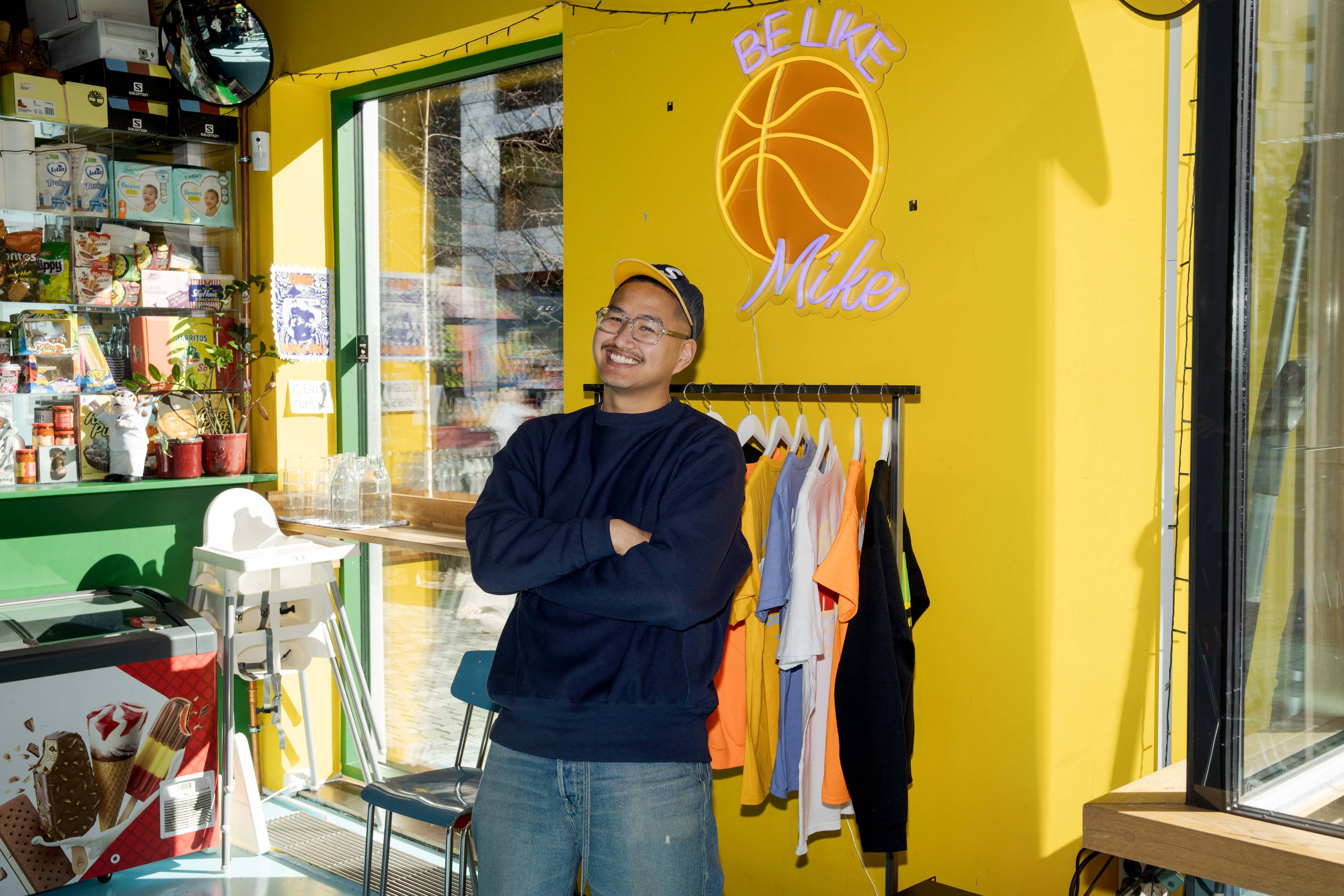 Mike poserer med armene i kryss og smiler. Bak han henger det noen tskjorter på et stativ og et basketballlys på den gule veggen.