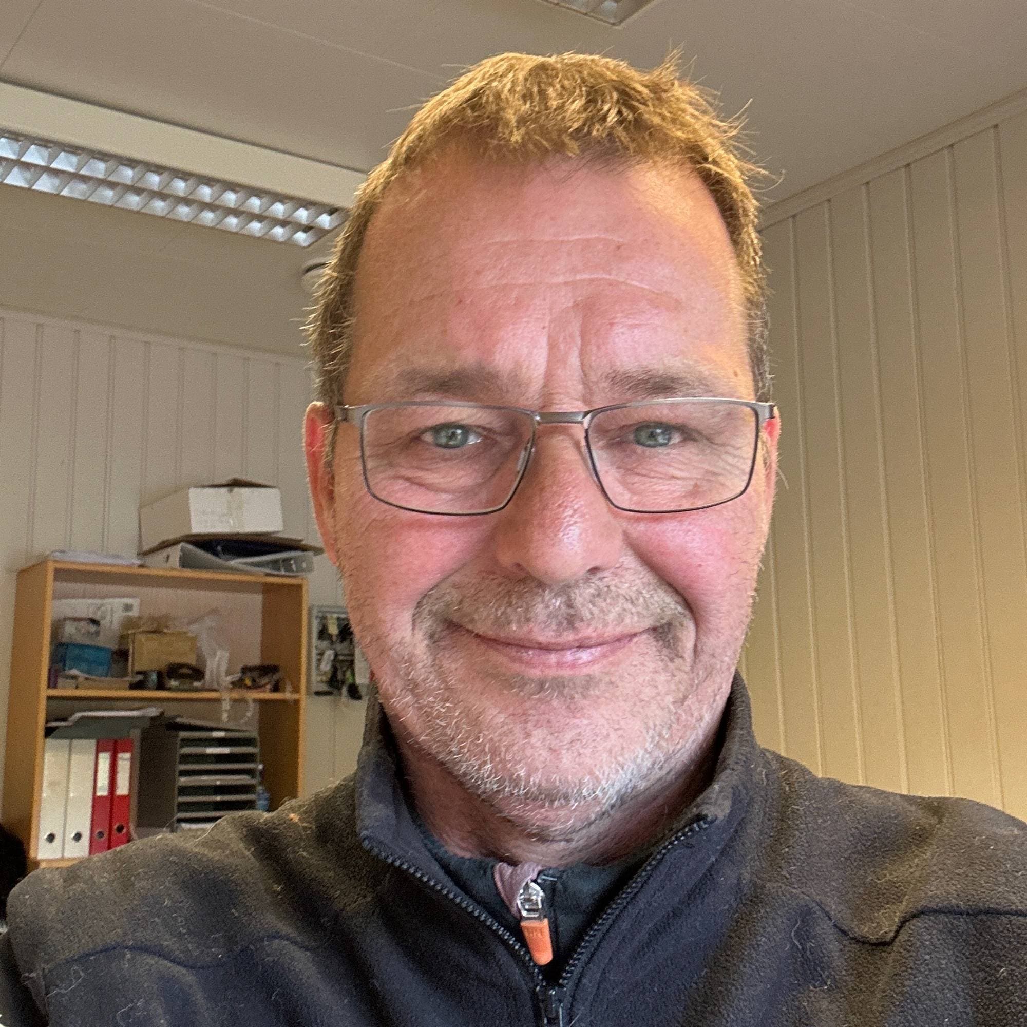 Portrett av en smilende mann med briller i en svart fleecejakke, tatt på et kontor. (Bildebeskrivelsen er laget av en KI-tjeneste)