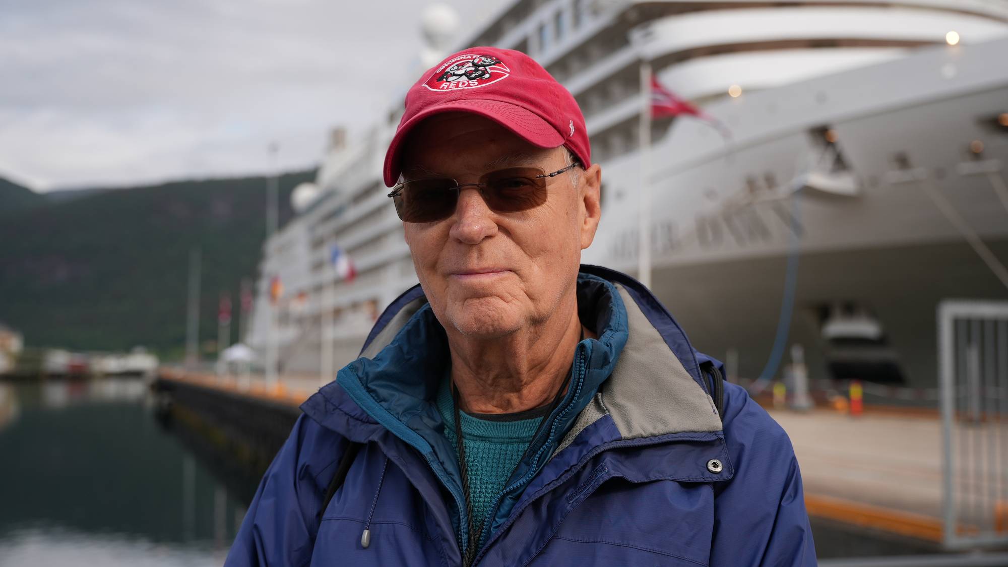En eldre mann med caps og solbriller står foran et stort cruiseskip. Mannen er kledd i en blå jakke og en grønn genser. Skipet er hvitt med flere etasjer og har flagg langs siden. I bakgrunnen sees en fjellkledd landmasse og en havnefront. (Bildebeskrivelsen er laget av en KI-tjeneste)