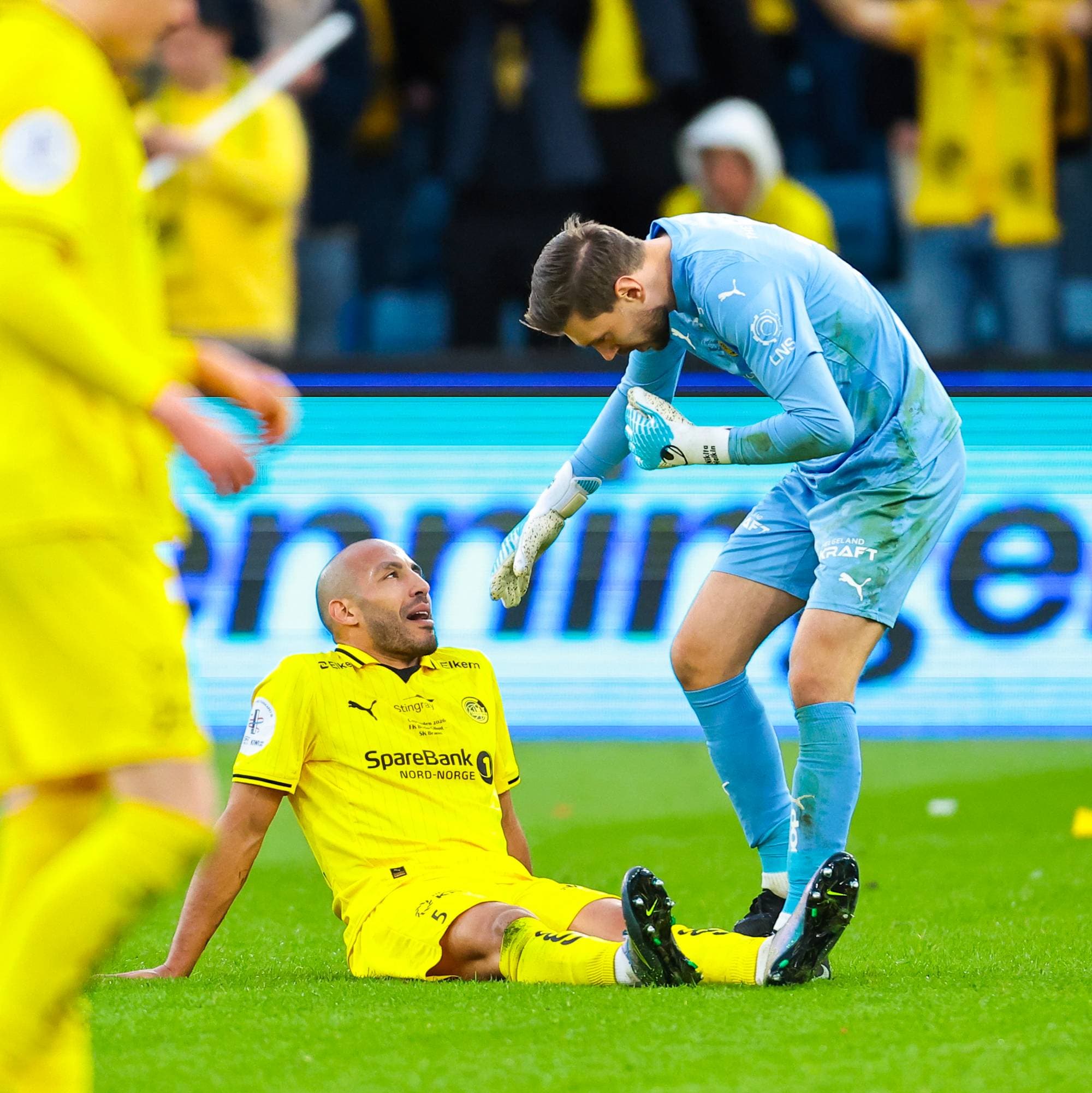 Keeper i lyseblått hjelper en sittende fotballspiller i gult opp fra gresset på en stadion. (Bildebeskrivelsen er laget av en KI-tjeneste)