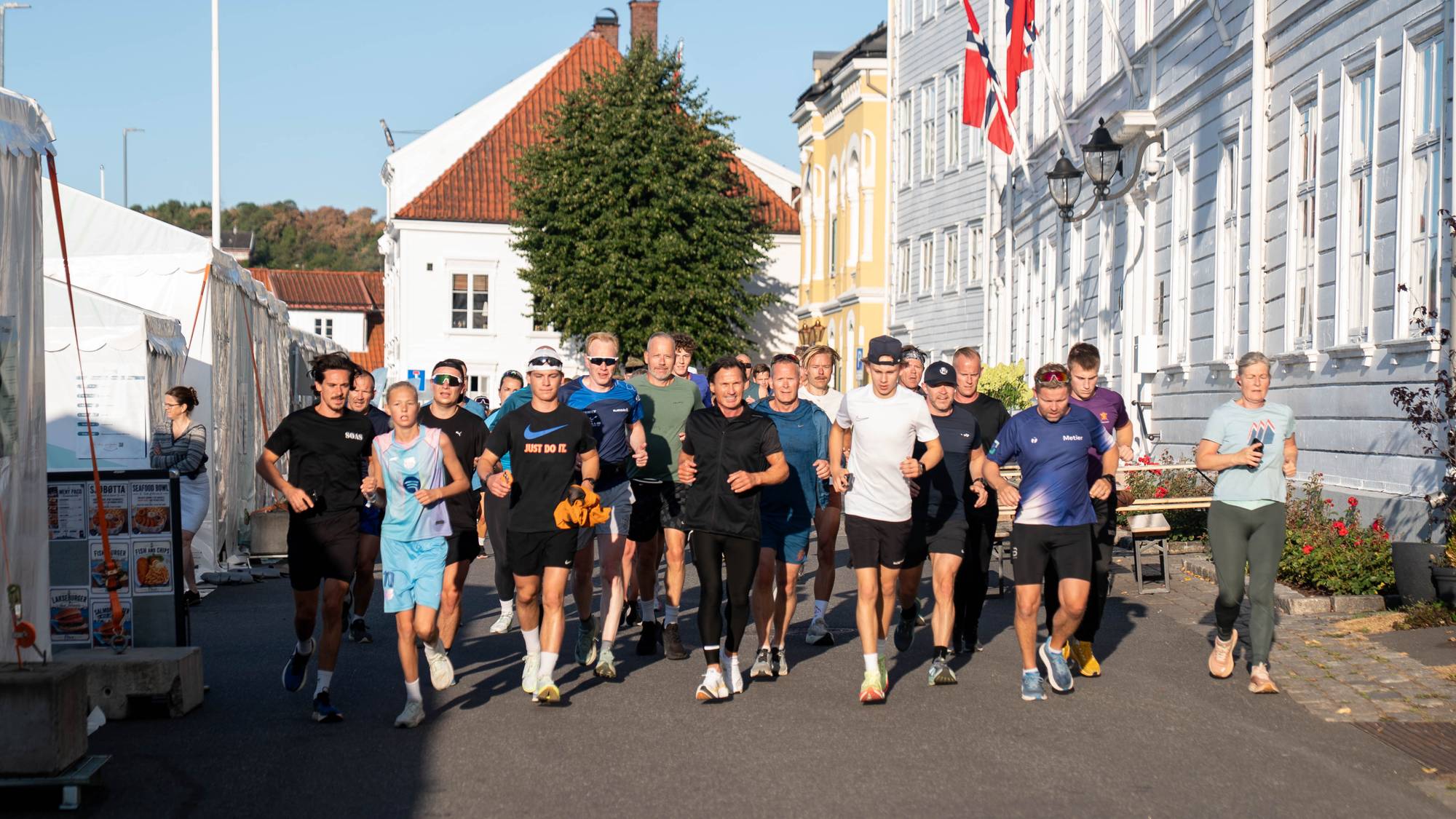 En gruppe mennesker løper sammen i en bygate med hvite trehus og norske flagg i bakgrunnen. 