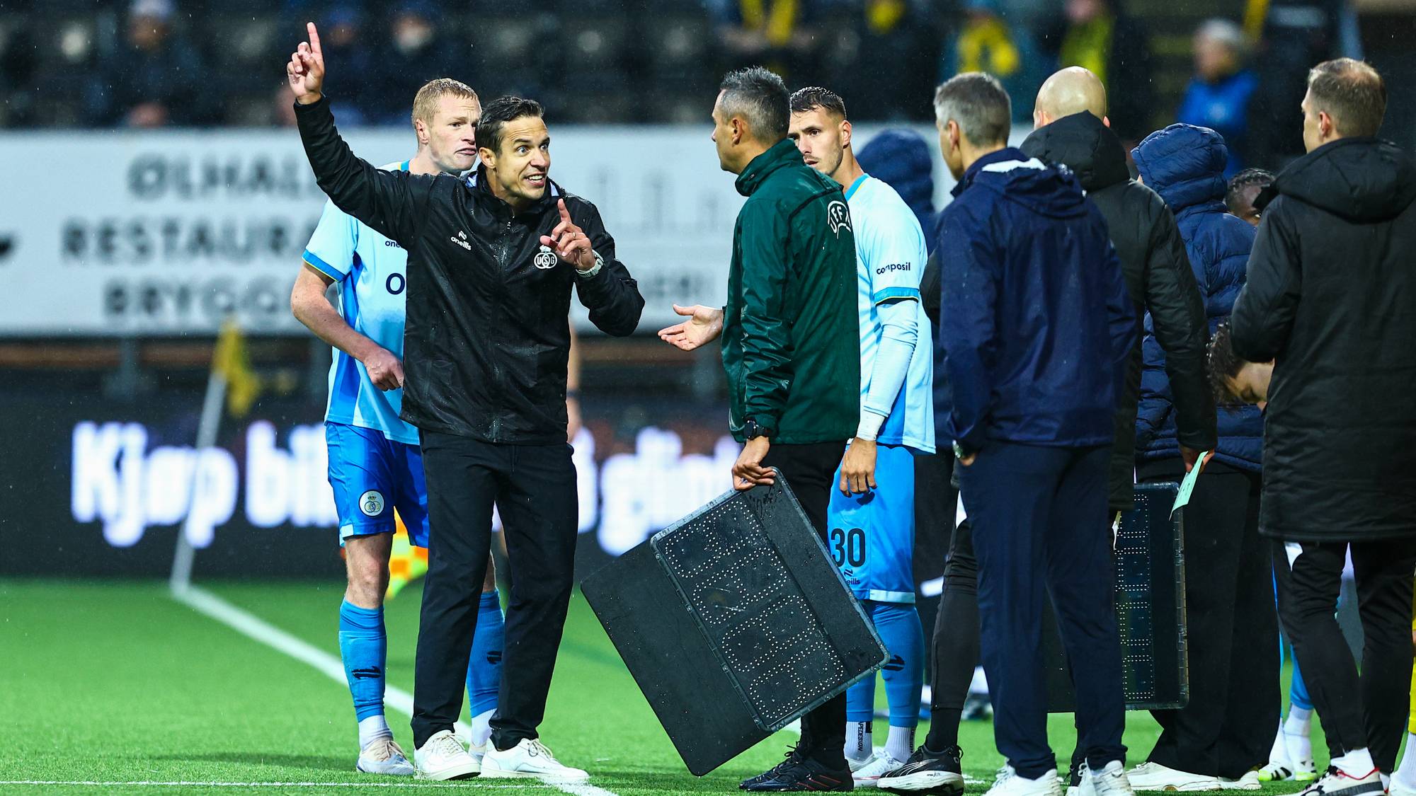 Fotballtrener gir instruksjoner til en spiller ved sidelinjen under et bytte med byttetavle. (Bildebeskrivelsen er laget av en KI-tjeneste)