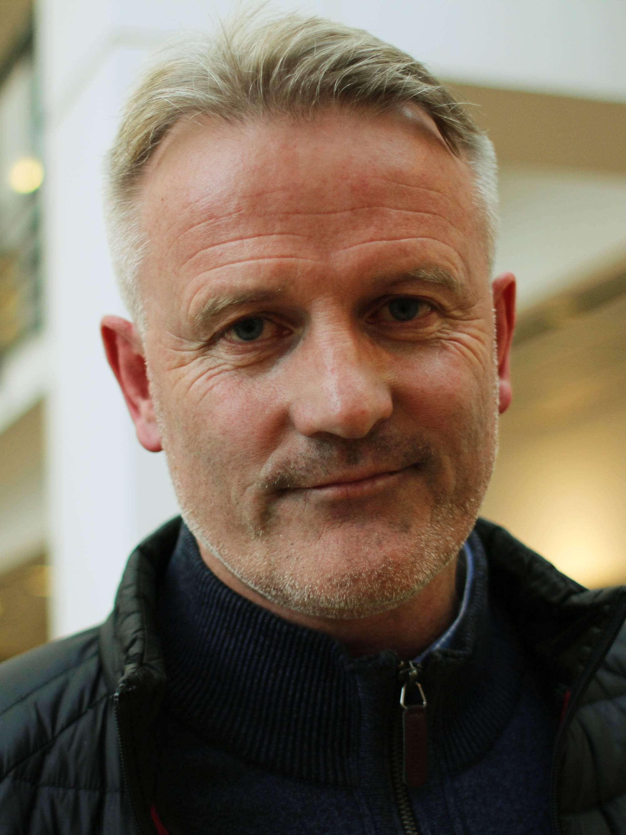 Mann med grått hår i svart jakke ser i kamera og smiler litt