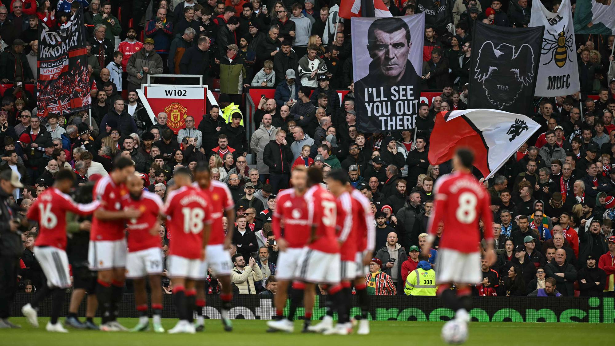 Manchester United-spillere på banen foran fans på tribunen med bannere og flagg. (Bildebeskrivelsen er laget av en KI-tjeneste)
